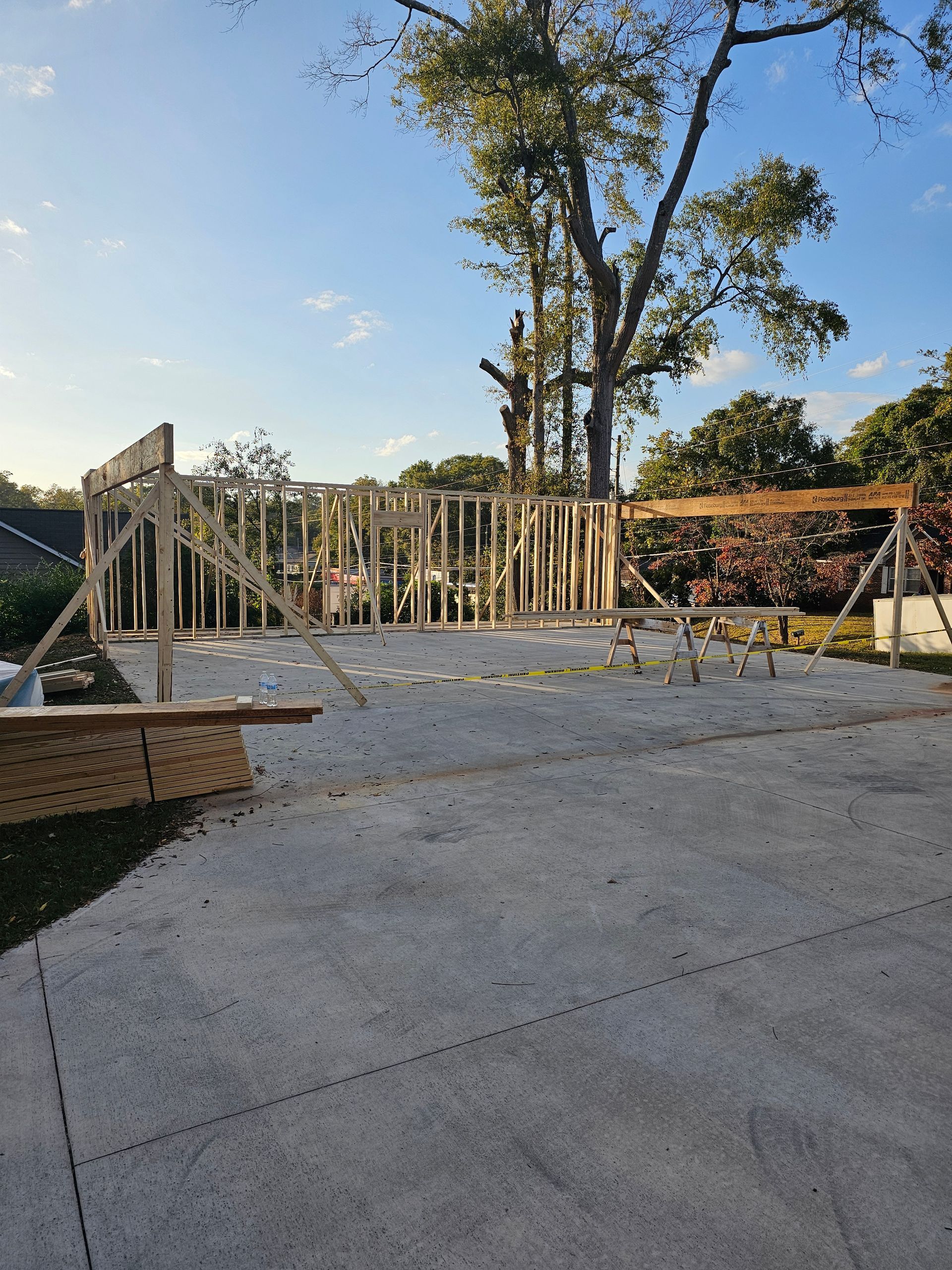 Wooden frame of a building under construction on a concrete slab, trees in background, sunny day.