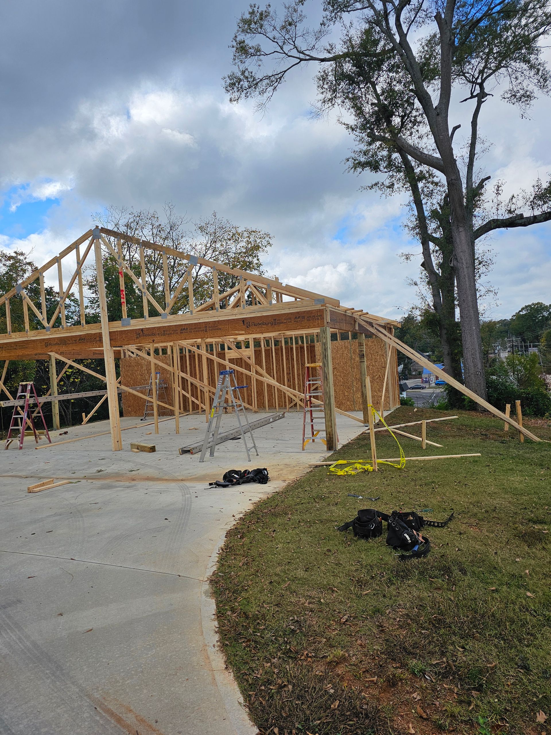 Wood frame structure under construction; concrete driveway and grass in foreground; cloudy sky.