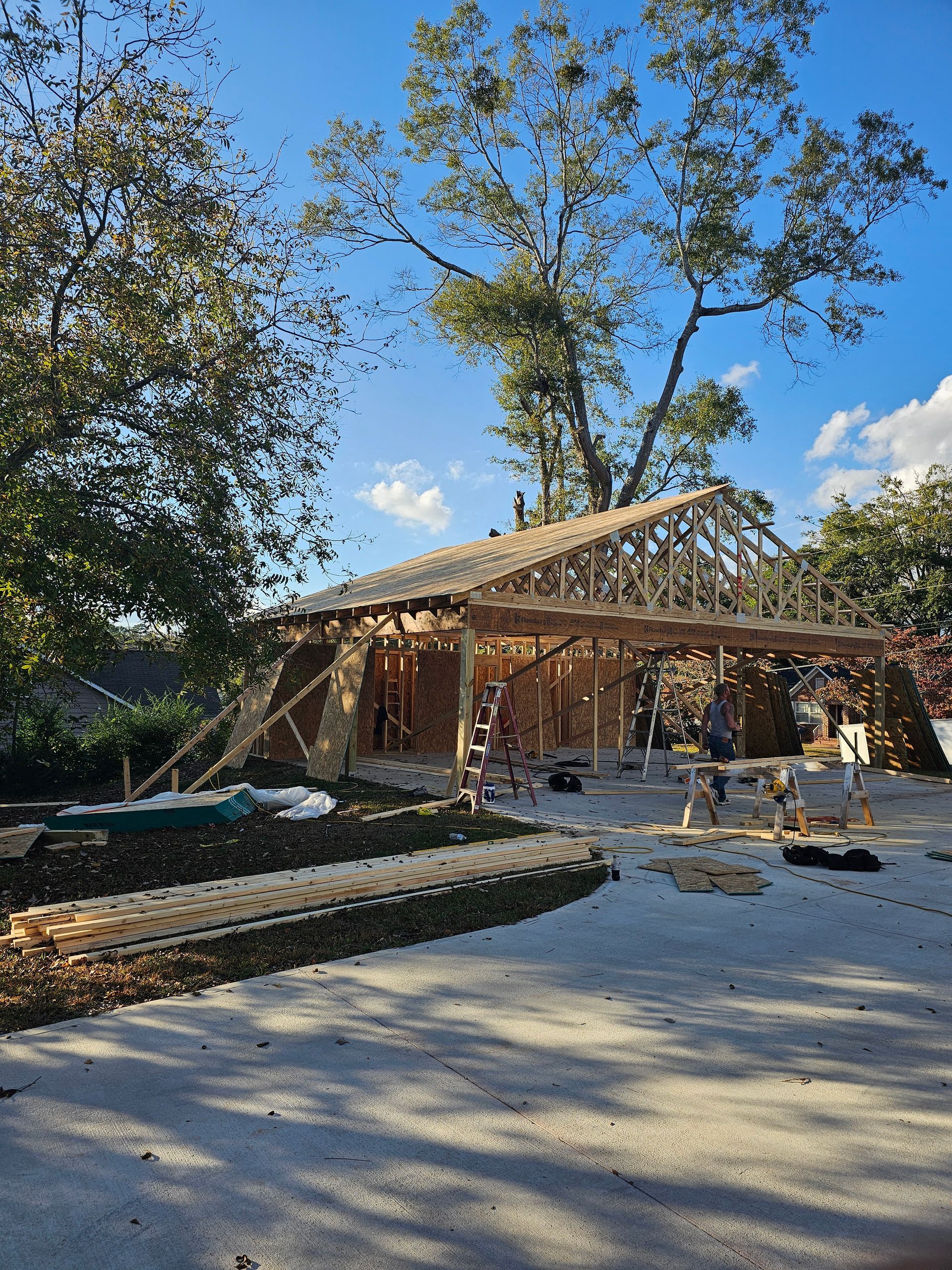 Construction of a wood-framed building; wooden beams and trusses visible; outdoors with trees and blue sky.