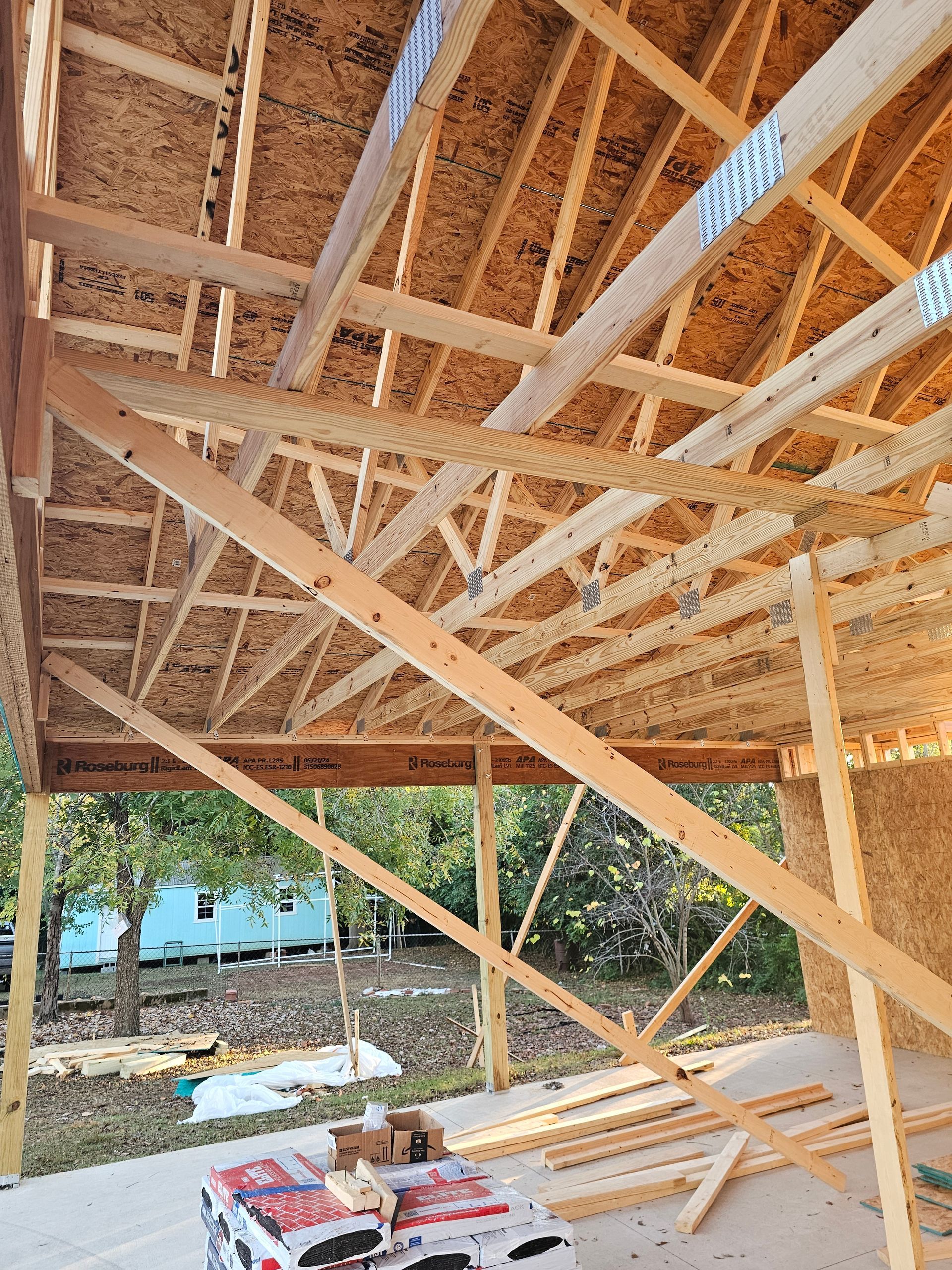 Construction of a wooden building's roof with exposed beams and support structures, viewed from underneath, outdoors.
