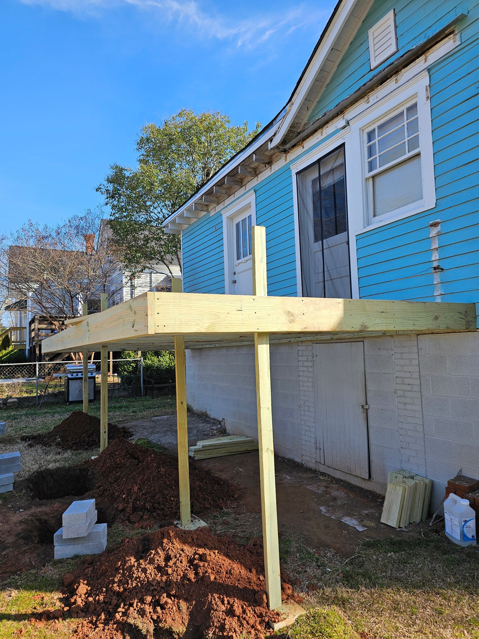 A deck under construction attached to a light blue house. Wooden beams, posts, and ground.