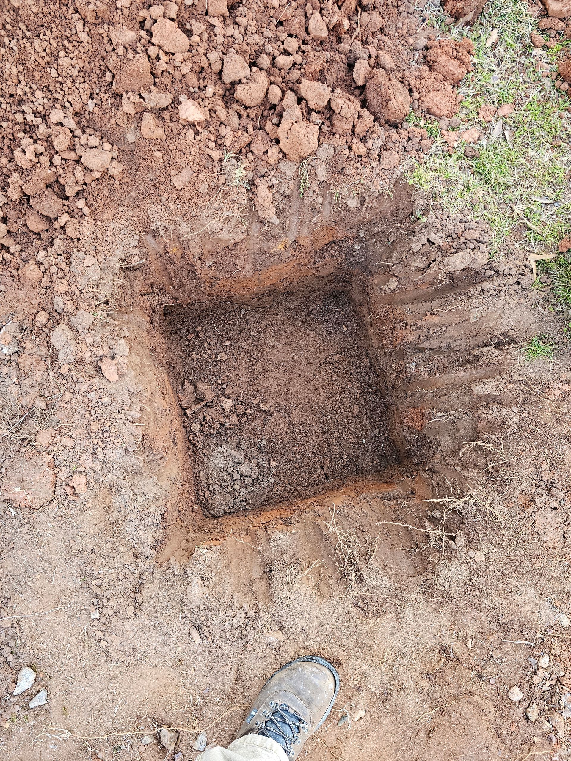 Square hole dug in reddish-brown dirt, with a foot visible near the bottom.
