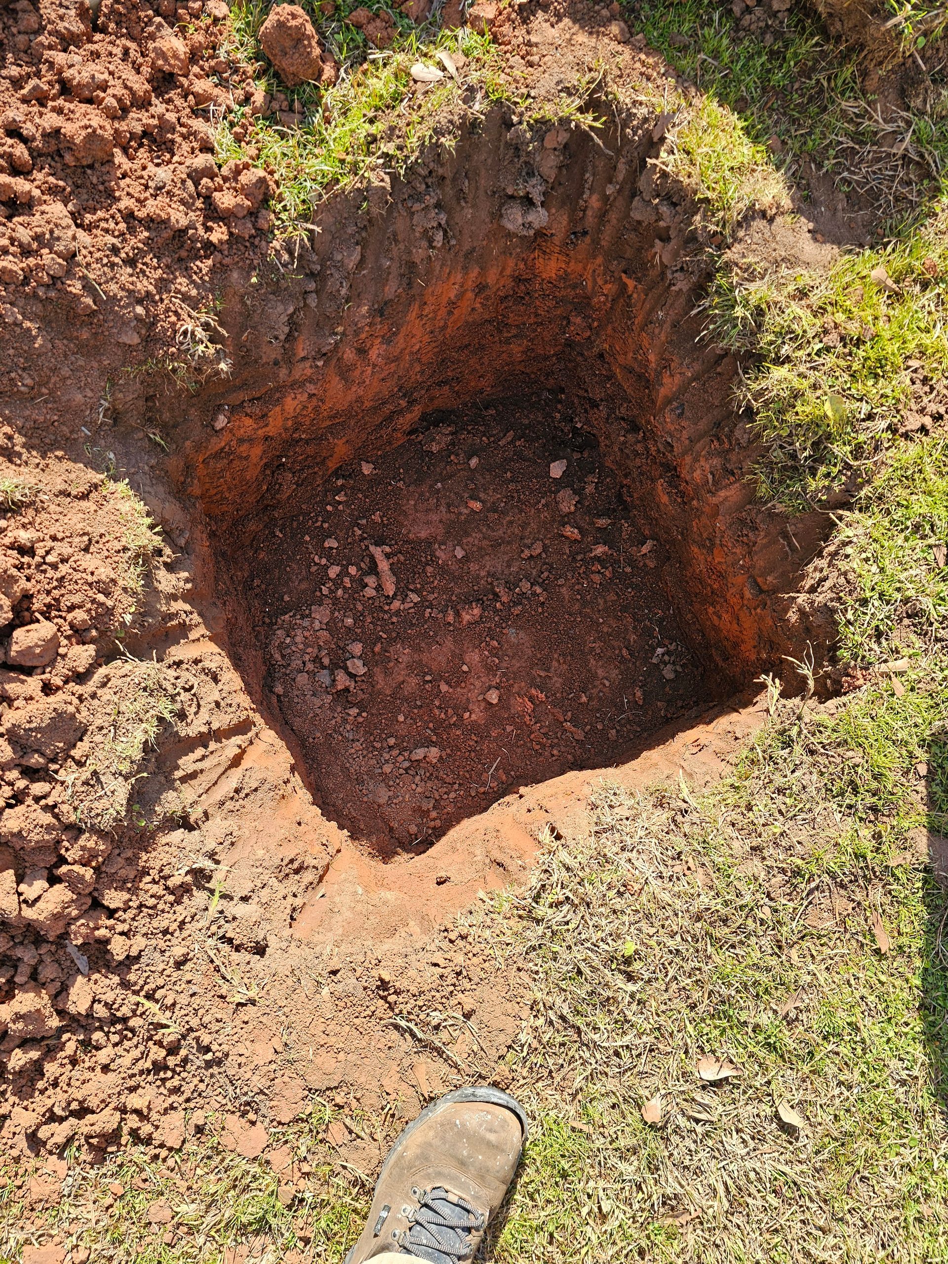 Square hole dug in reddish-brown soil, surrounded by grass, with a foot visible at the bottom.