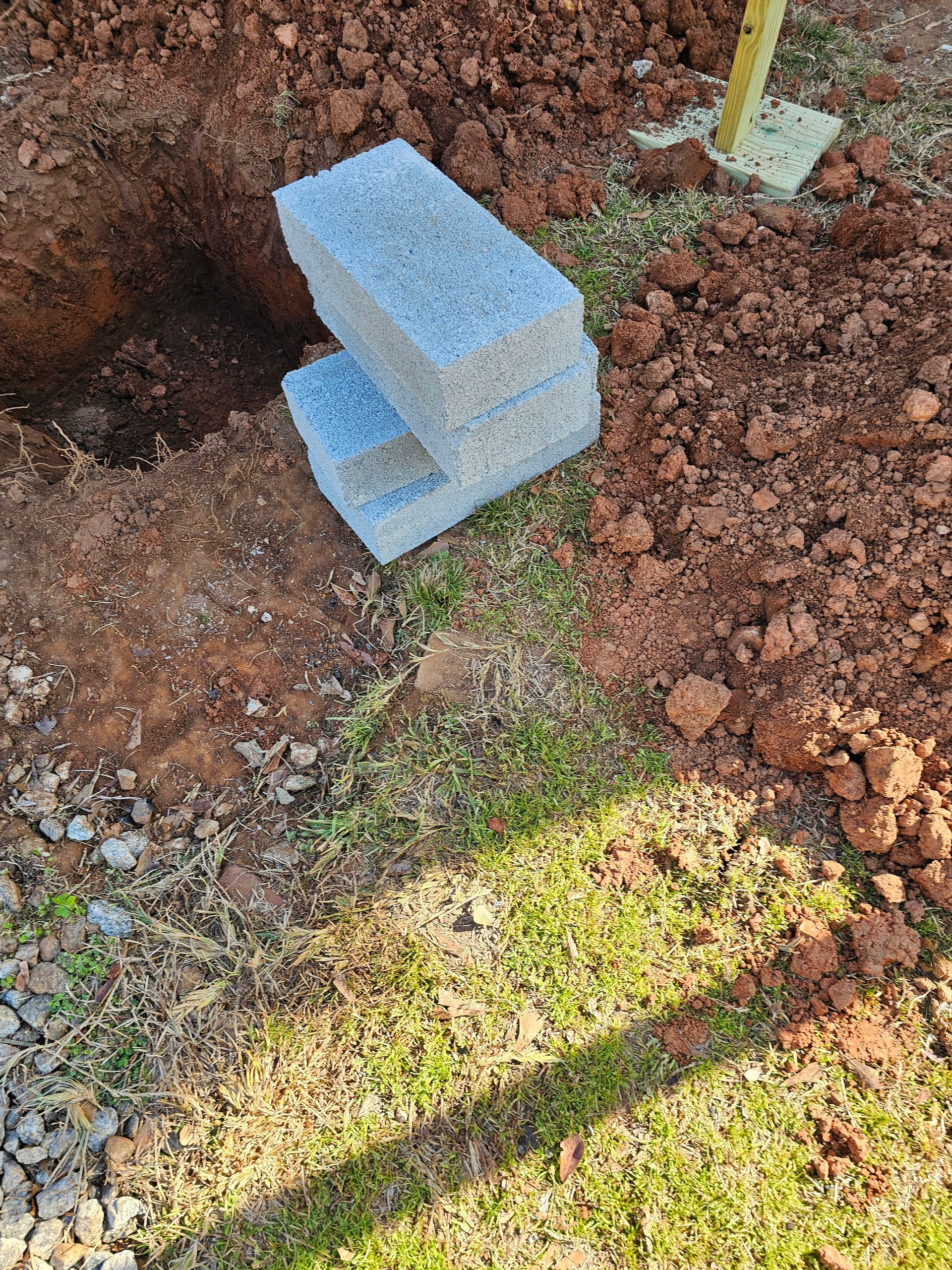 Pile of gray cinder blocks next to a dug hole in reddish-brown dirt. Grass in foreground.