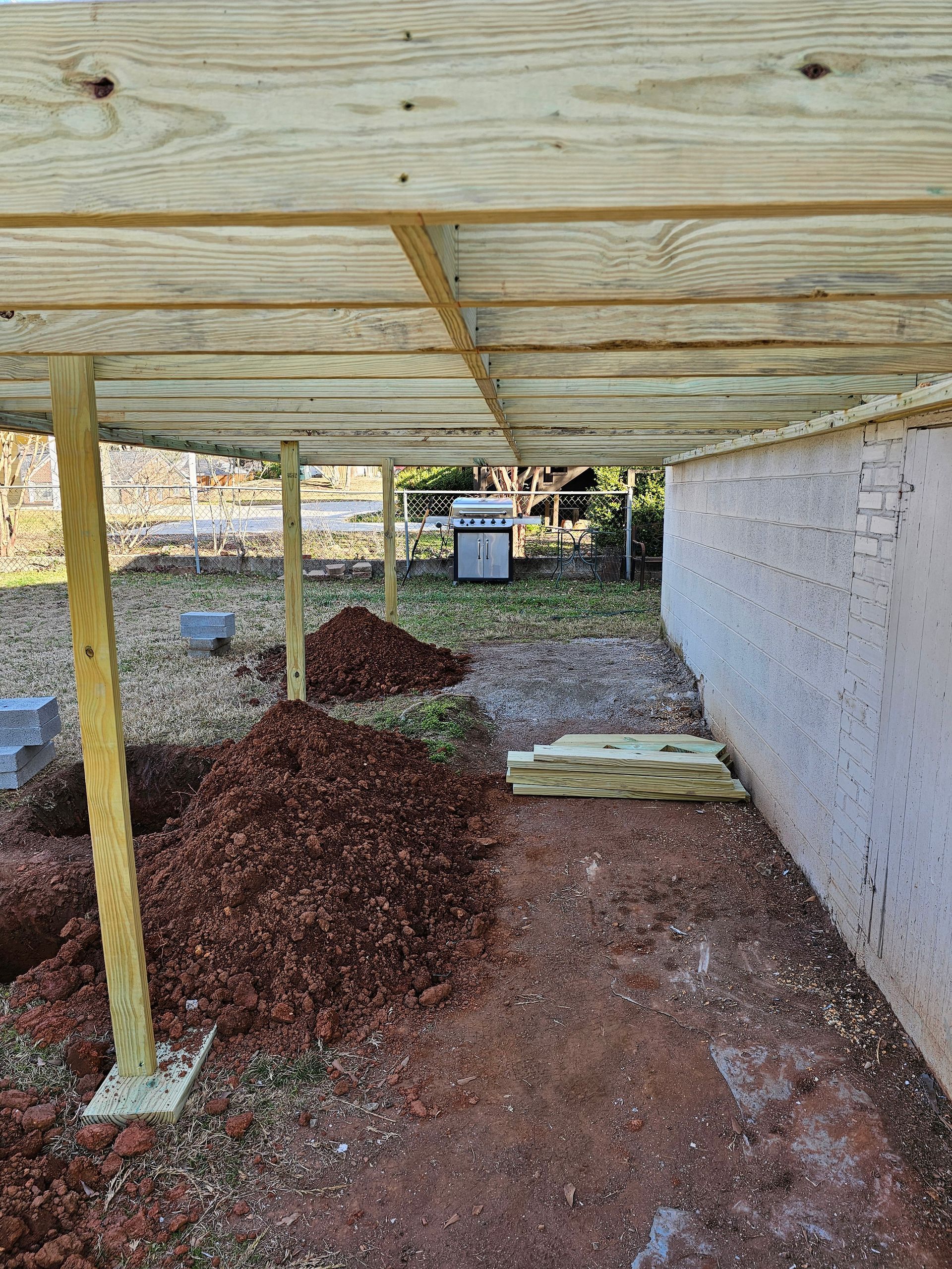 Under-construction deck with wooden supports, dirt piles, and a white brick wall.