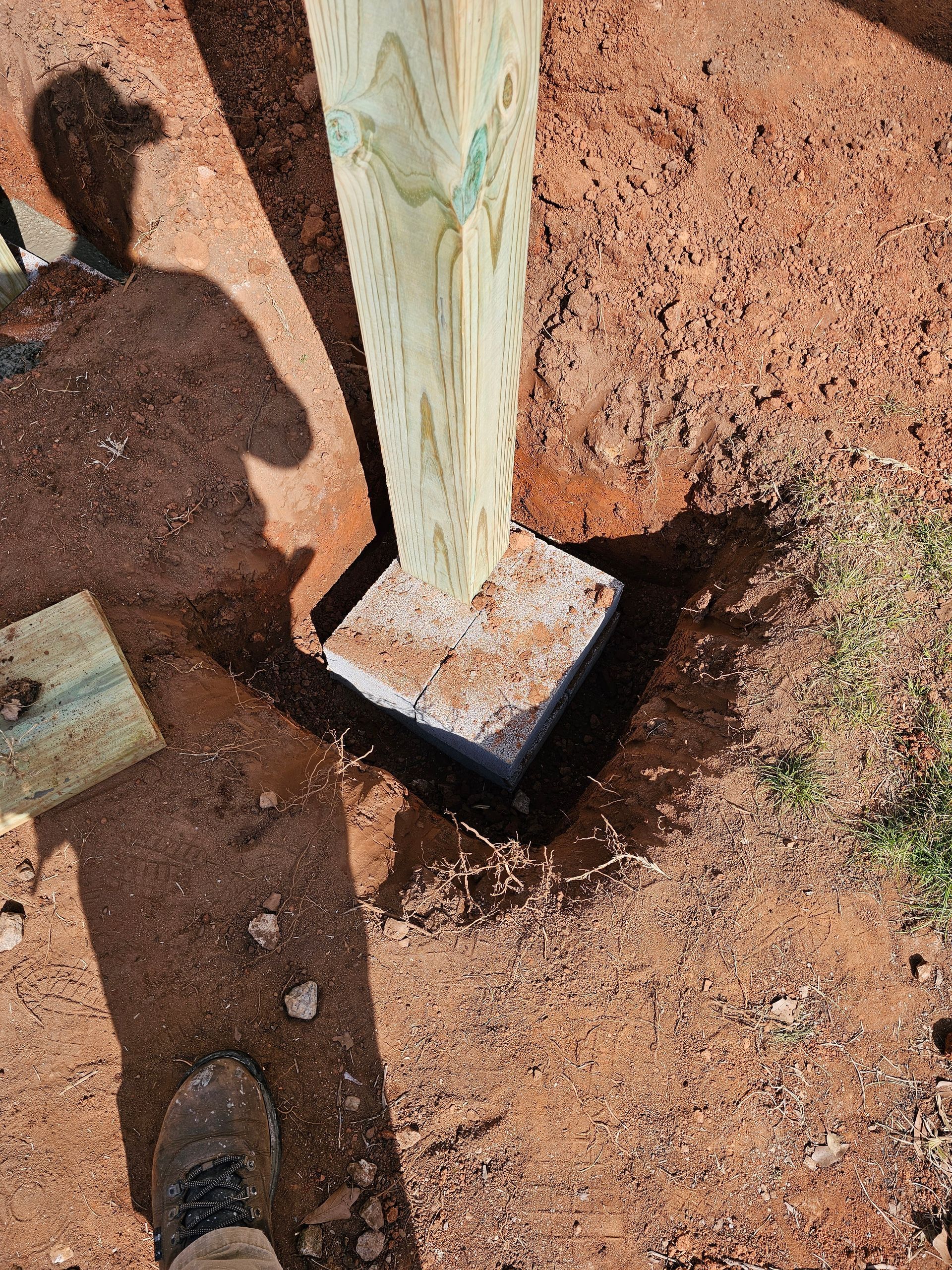 Wooden post set in a concrete base, in dirt; a shadow of a person.