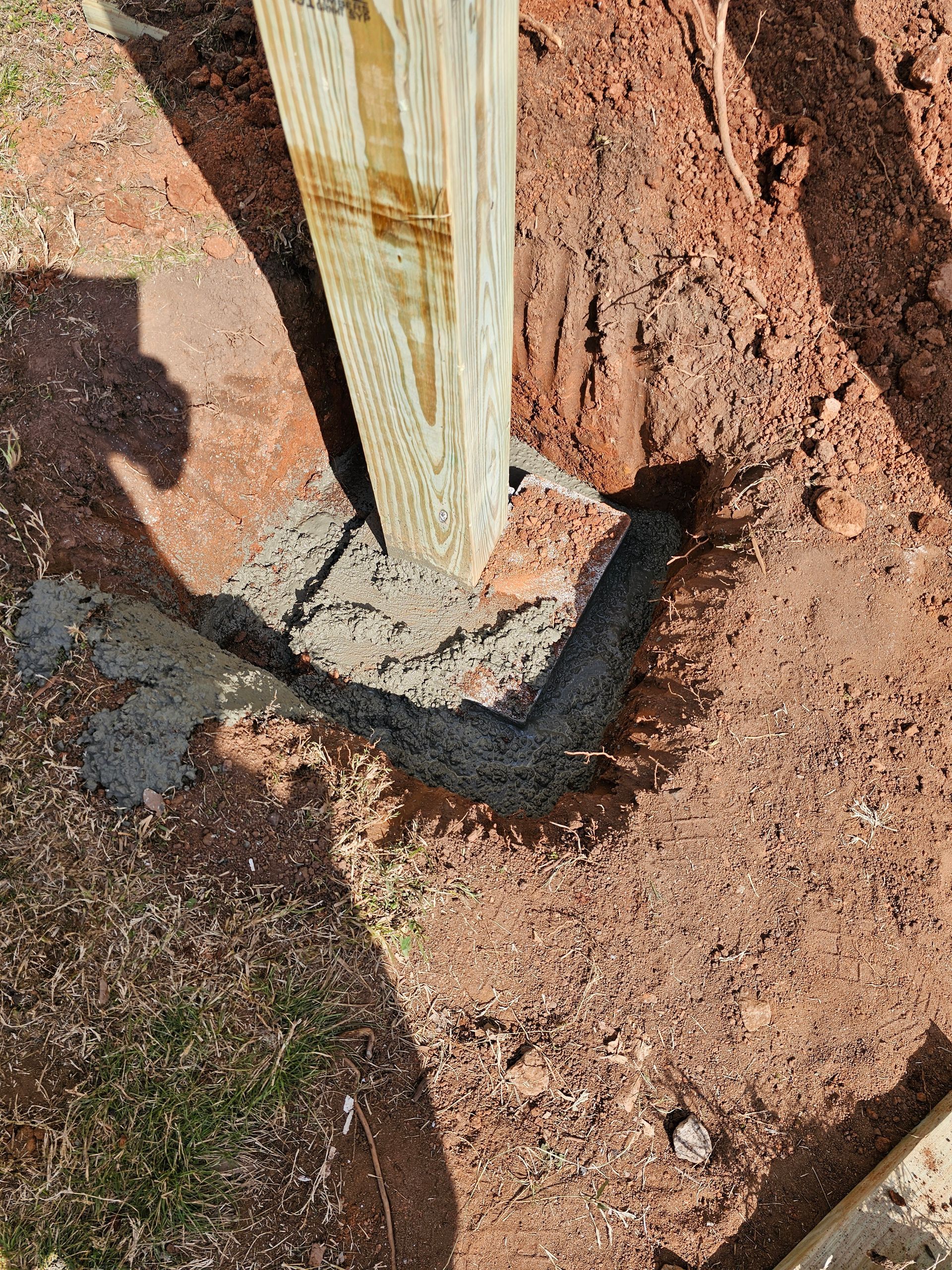 Wooden post set in wet concrete in a dirt hole. Red-brown soil surrounds the base.