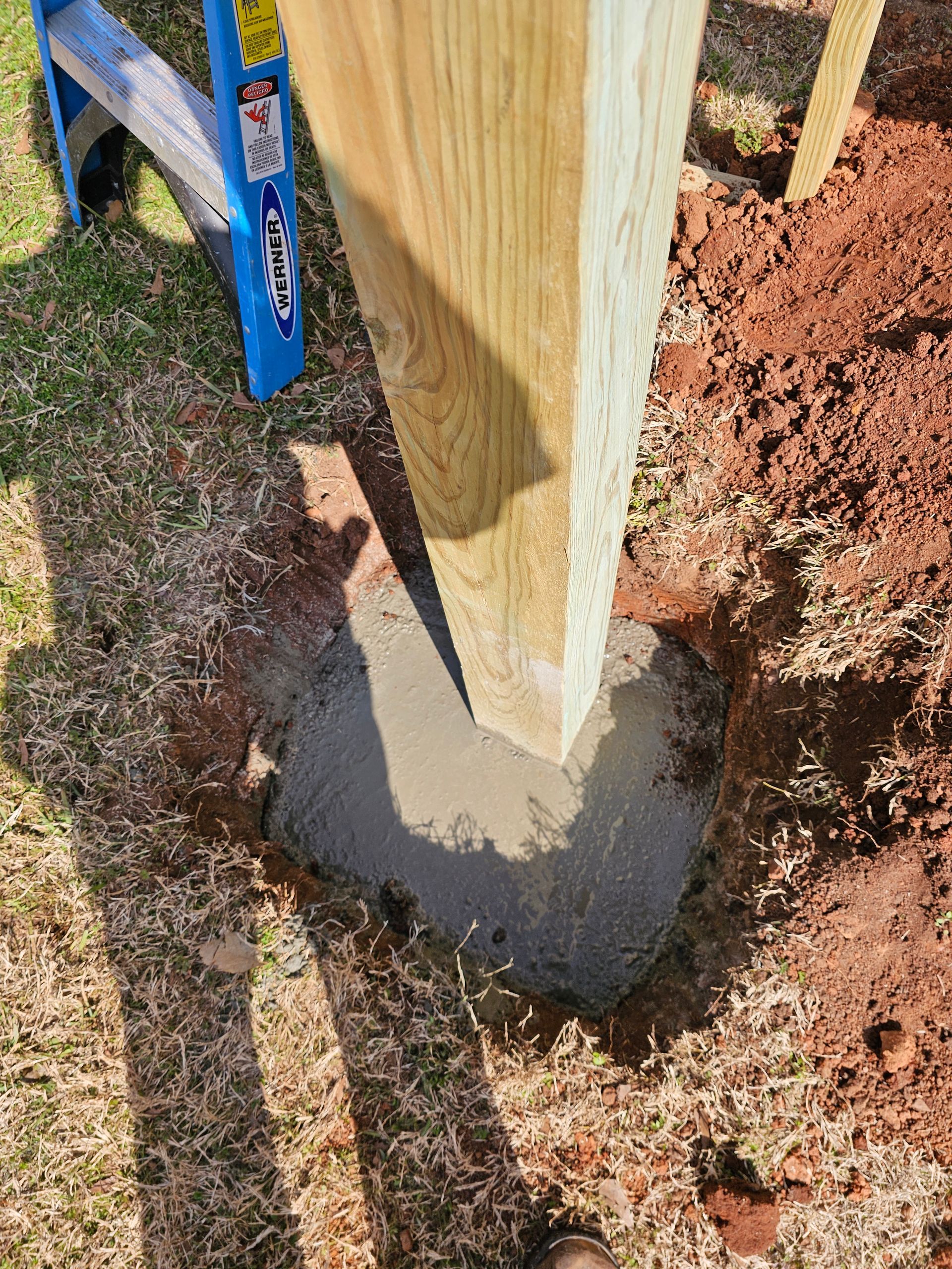 Wooden post set in wet concrete, in a small hole. A blue ladder and brown dirt are visible in the outdoor setting.