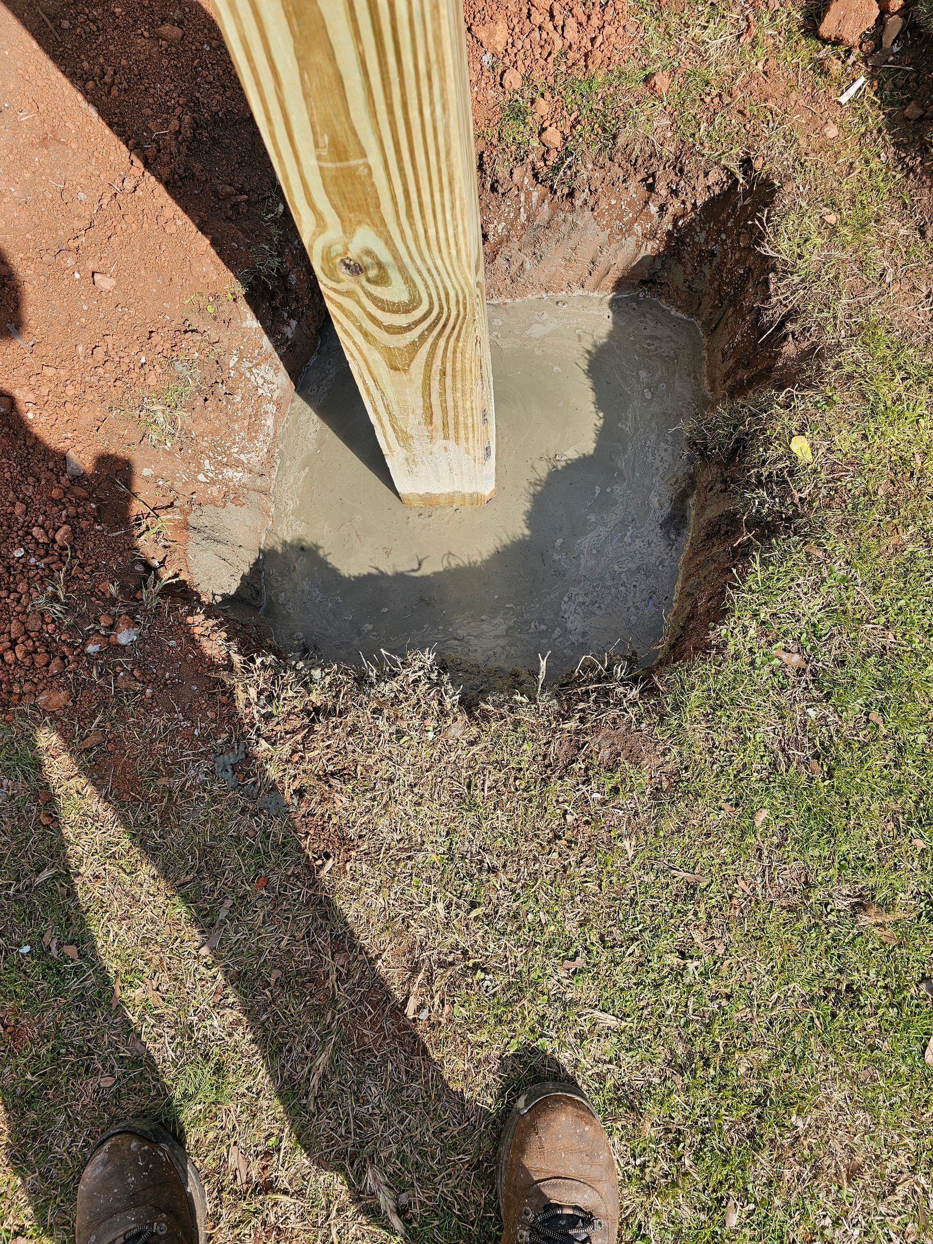 Wooden post set in wet concrete in a hole in the ground, surrounded by grass and dirt.
