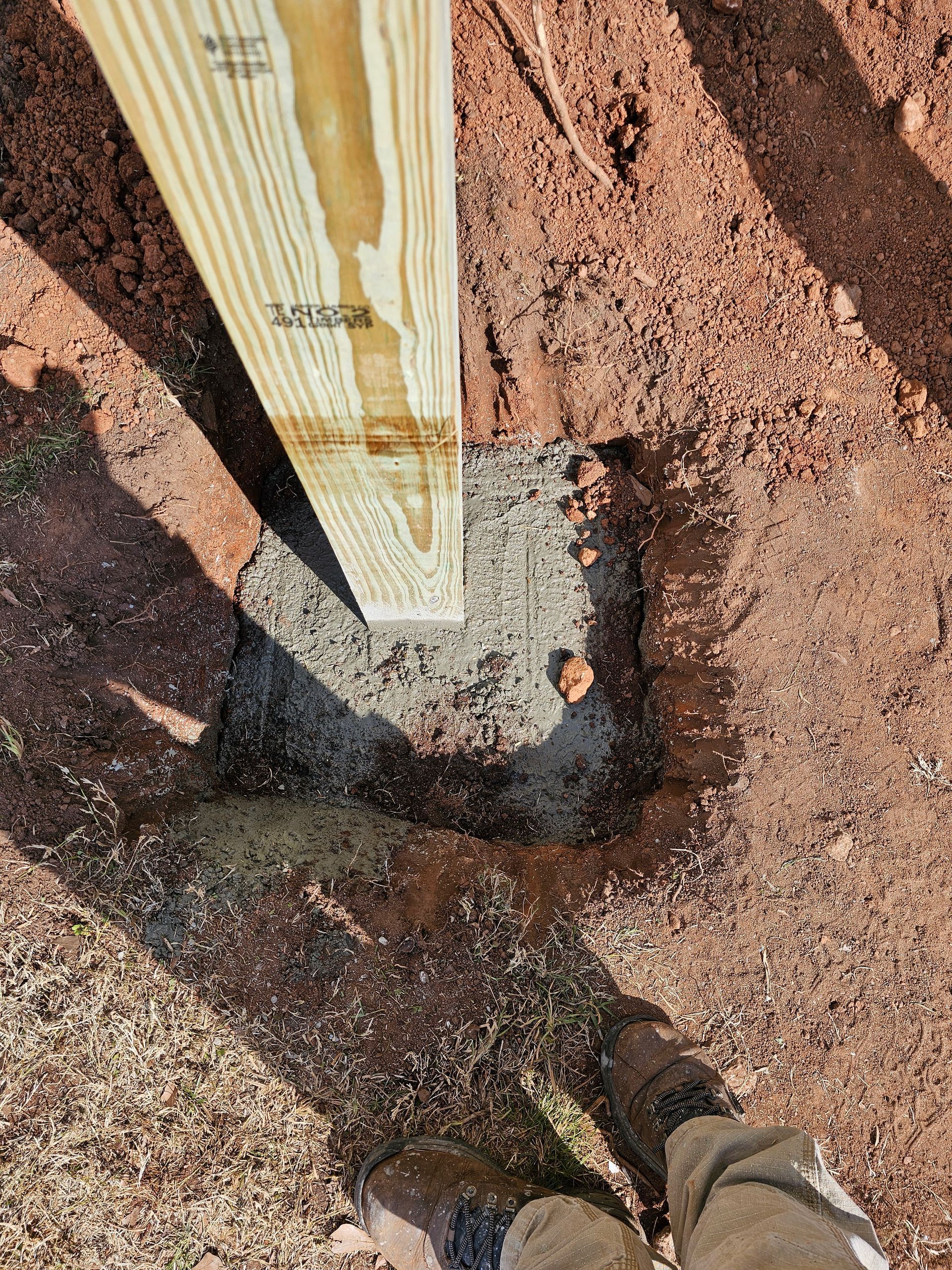 Wooden post set in concrete in a square hole in reddish-brown soil. A person's brown boot is visible.