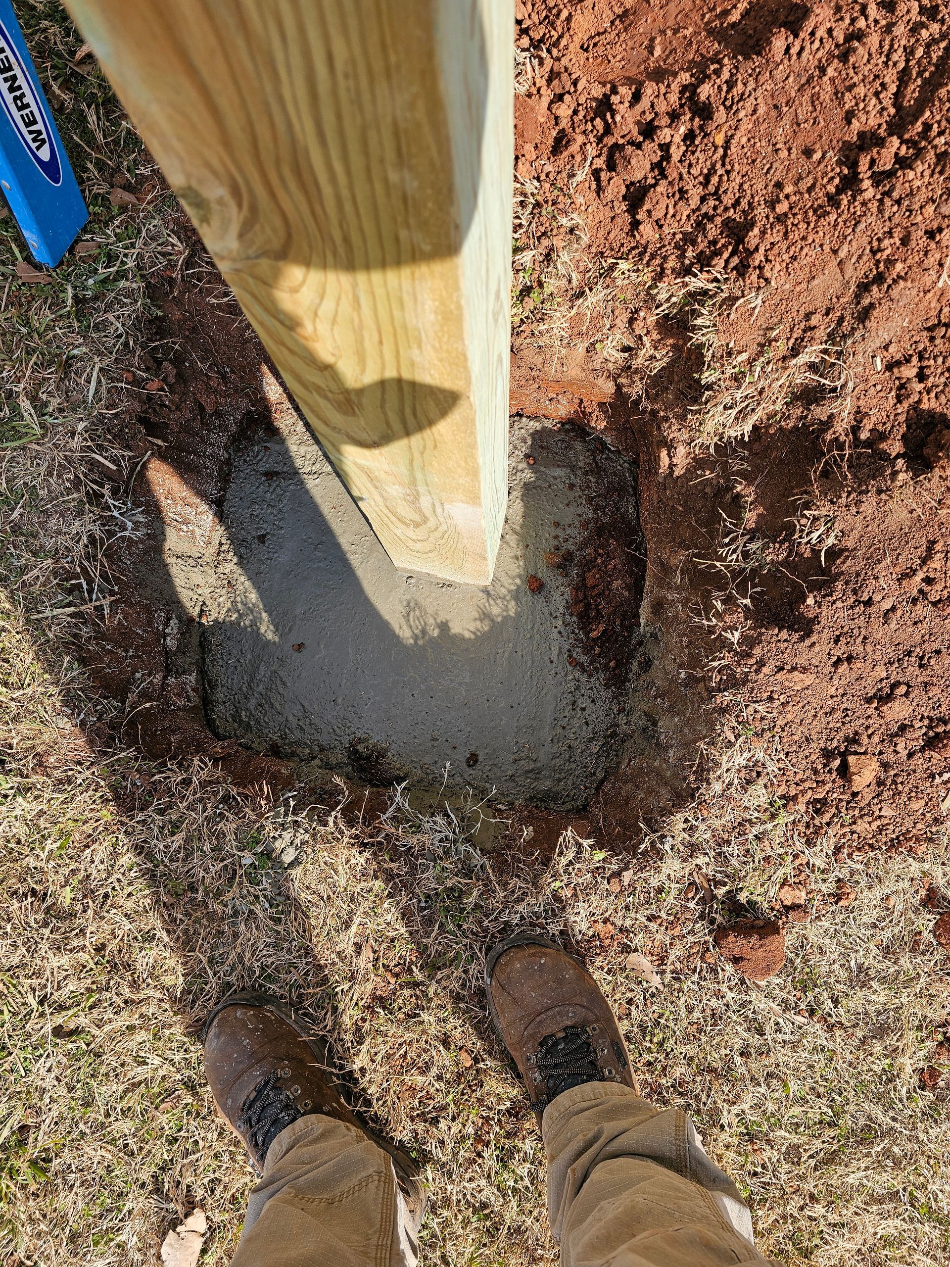 Person standing near a wooden post set in concrete in a hole in the ground.