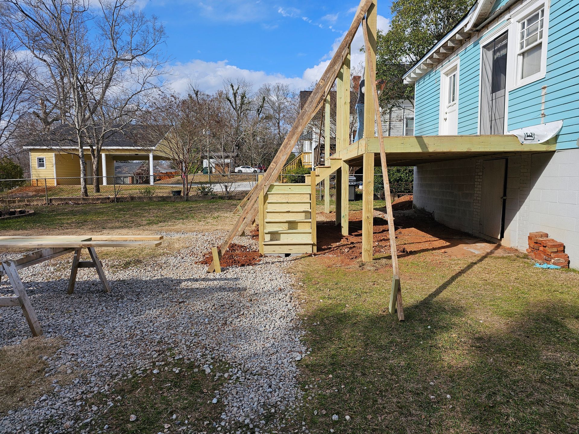 A partially built wooden deck with stairs next to a blue house, construction materials in yard.
