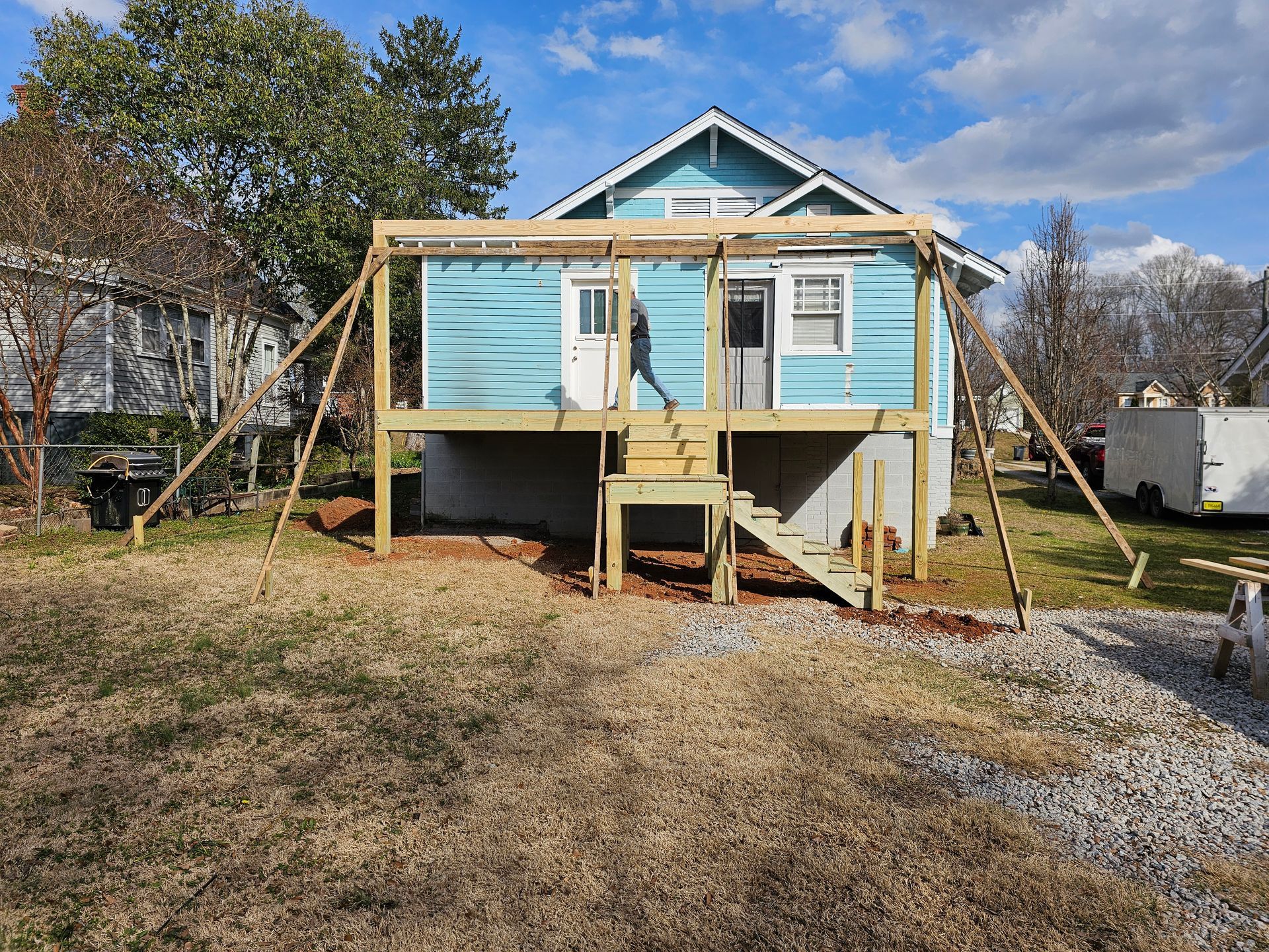 A blue house with a wooden deck under construction; wooden supports stabilize the deck on a grassy lot.