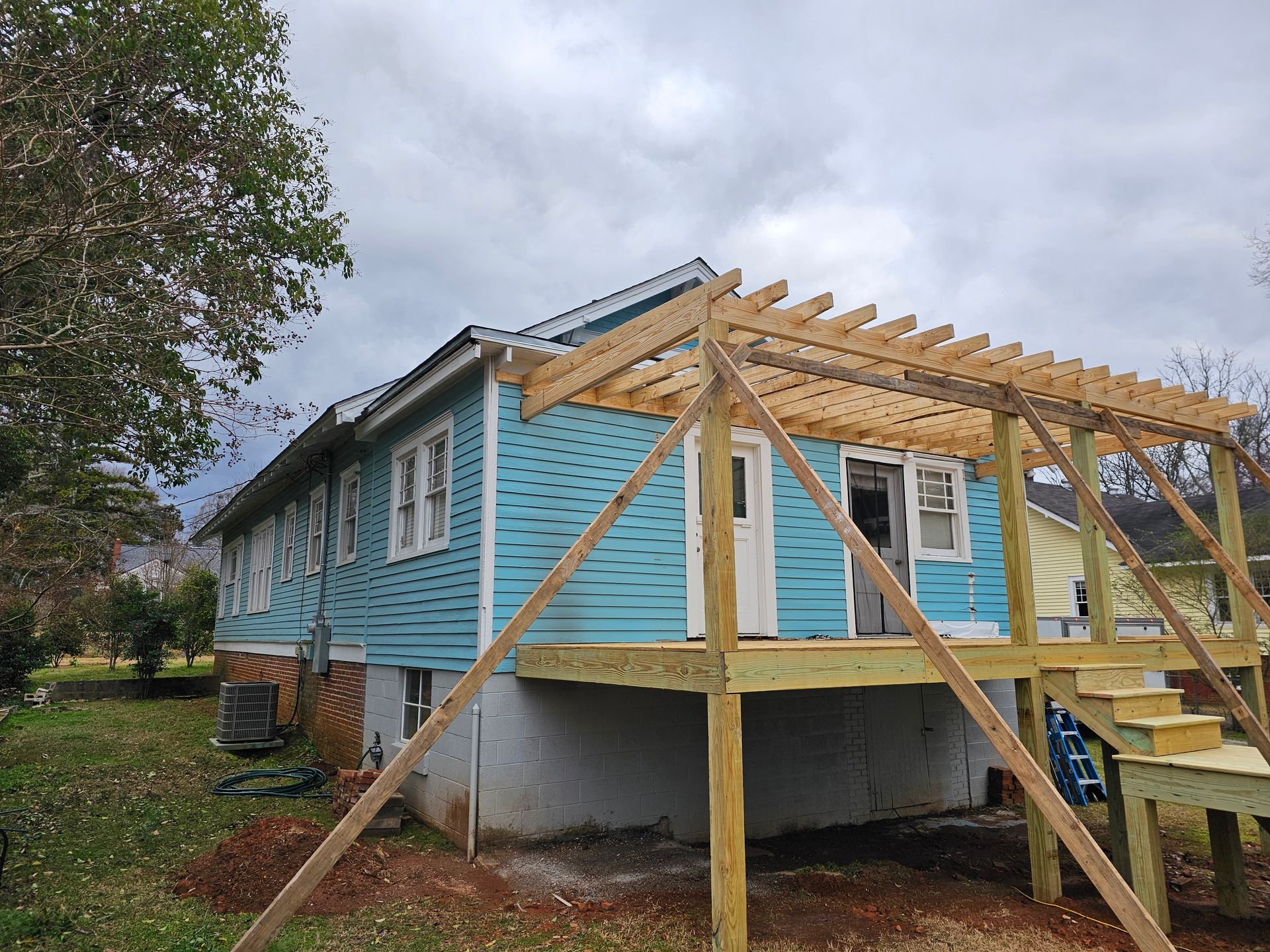 Blue house with wooden deck, pergola, and stairs under cloudy sky.