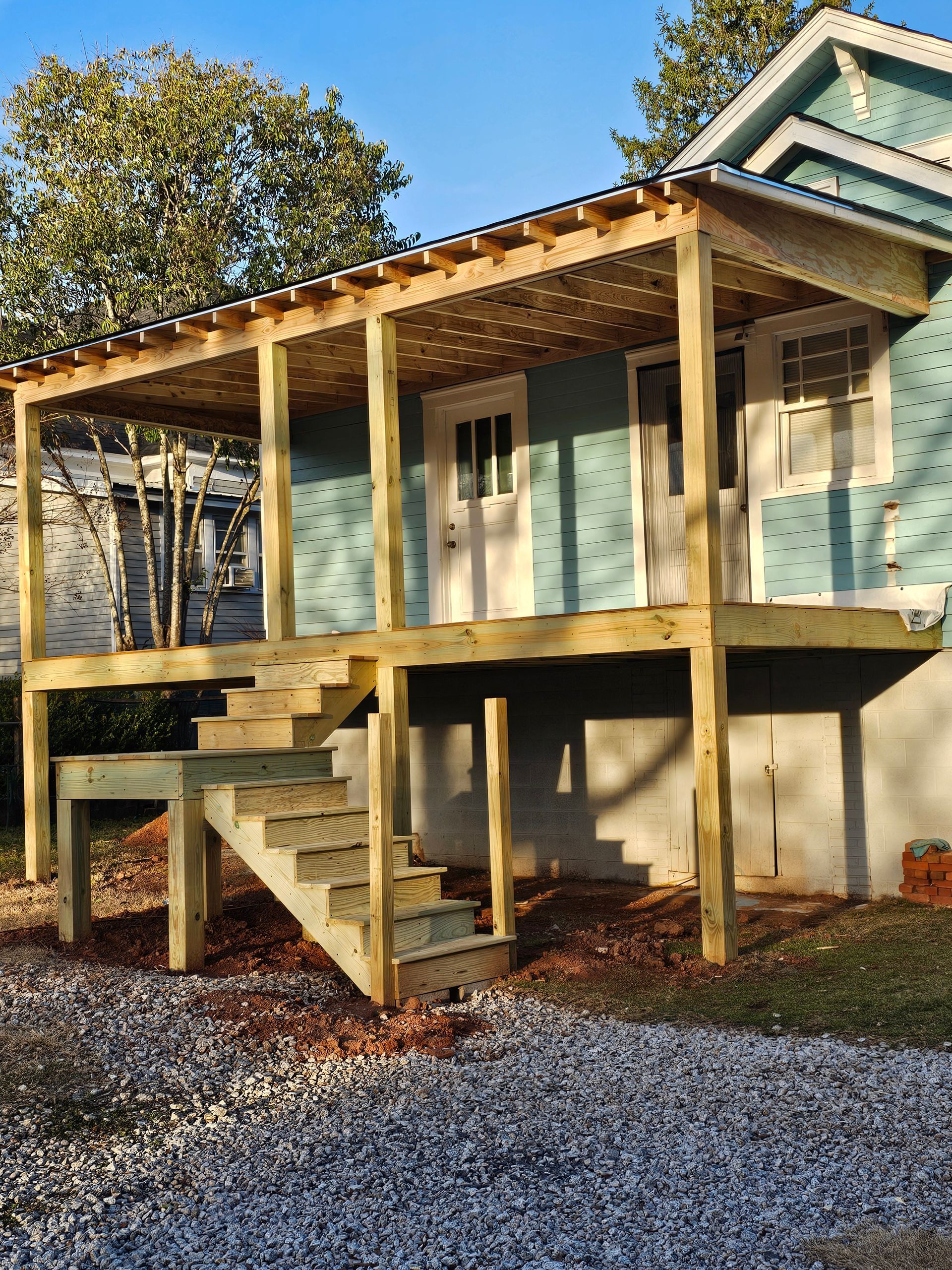 Wooden deck and pergola attached to a blue house with a set of stairs leading to a gravel yard.