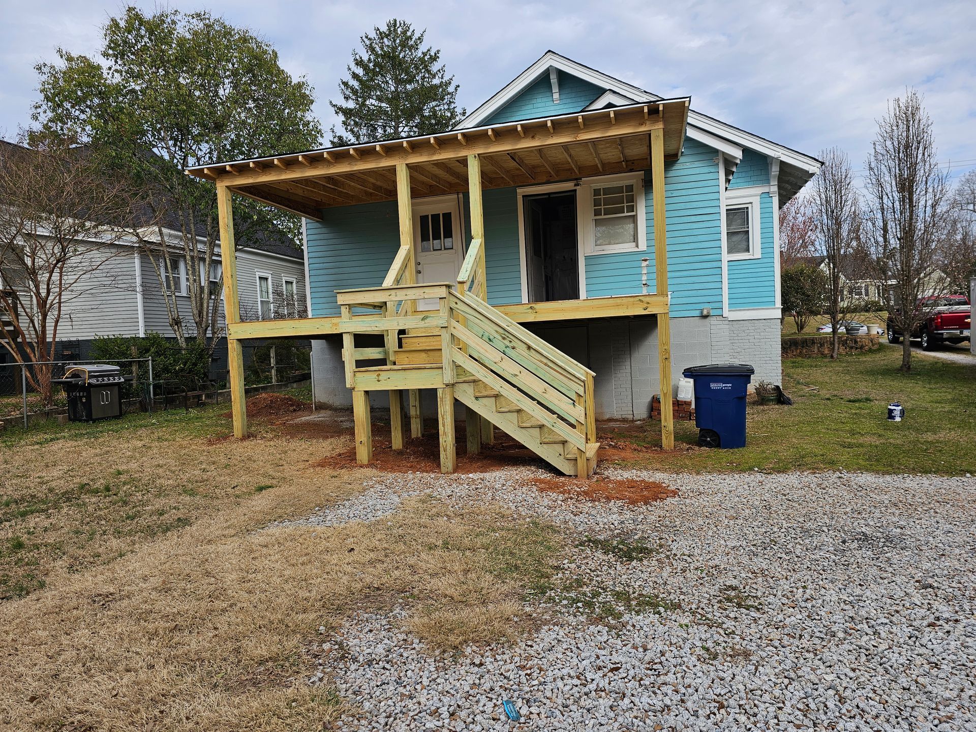 Small blue house with a wooden porch and stairs, in a yard with gravel.