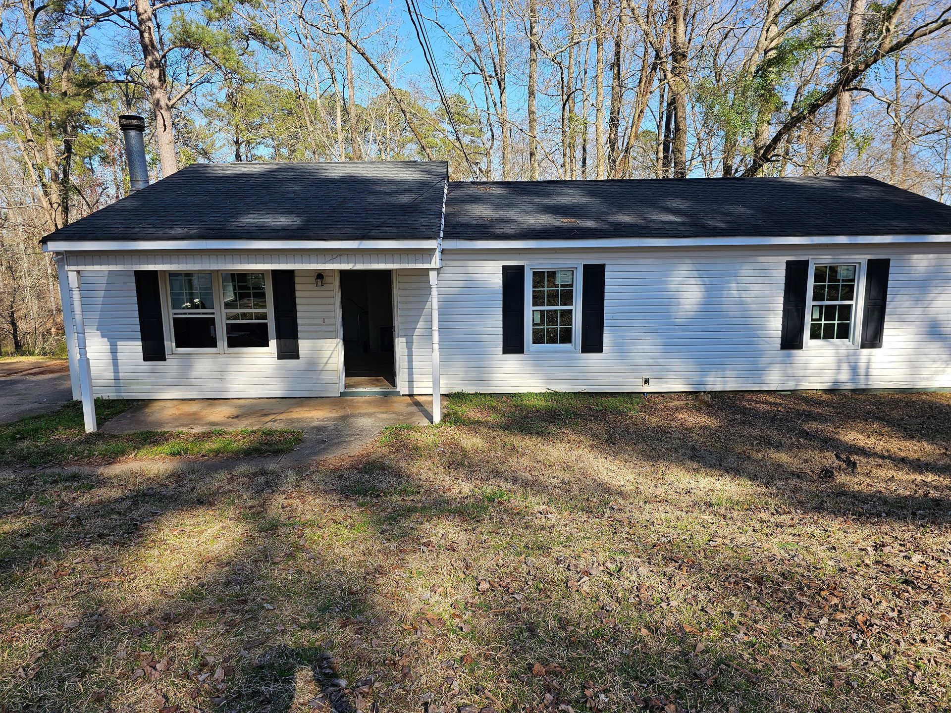 White single-story house with black shutters, on a grassy lot, trees in the background under a blue sky.