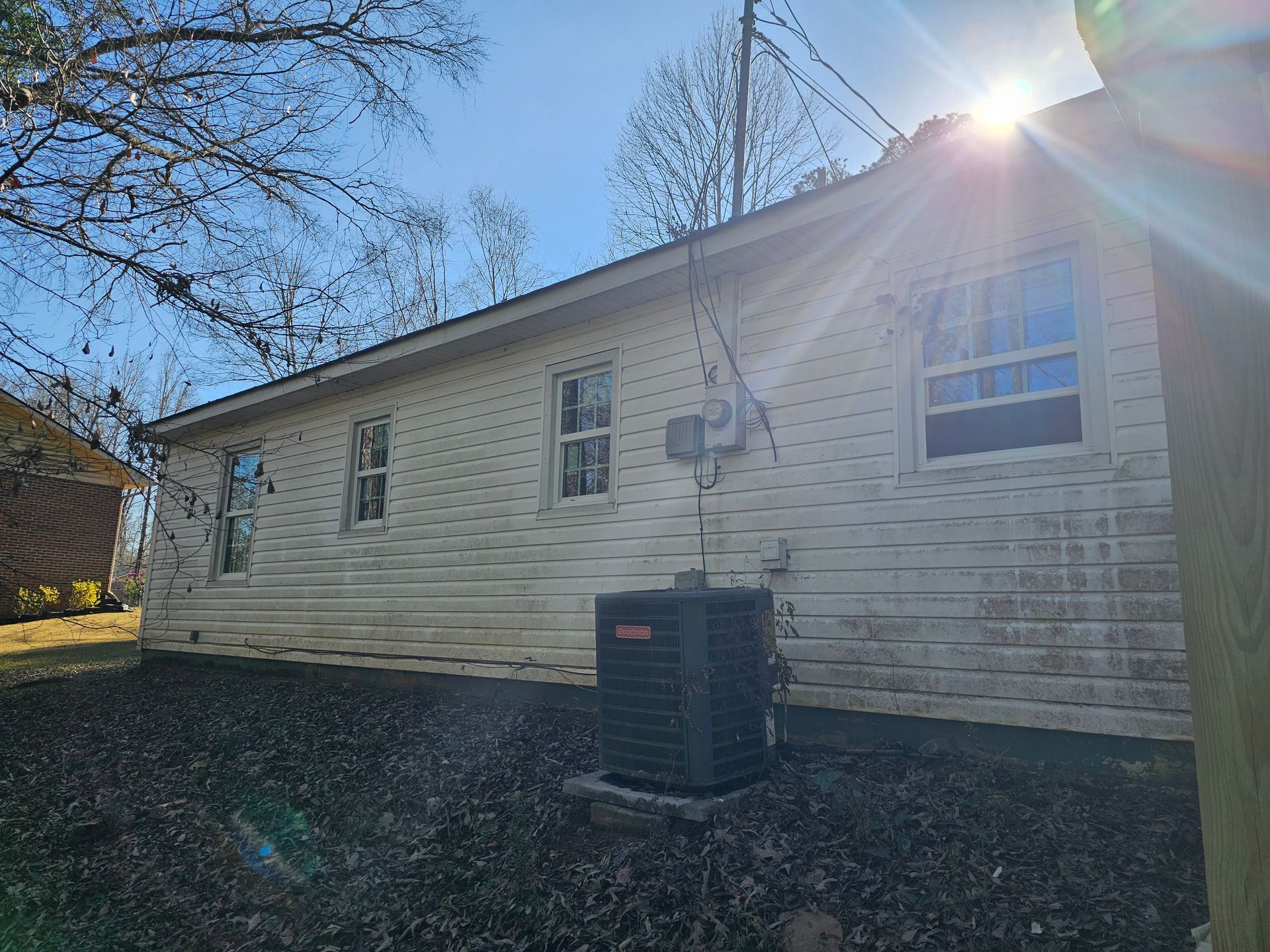 Side of a white house with three windows, an air conditioner, and the sun shining.