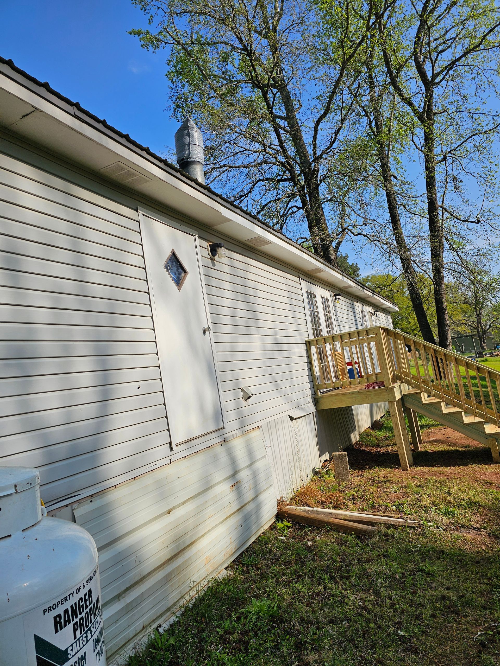 Side view of a light gray house with a wooden deck and a propane tank in a grassy yard.