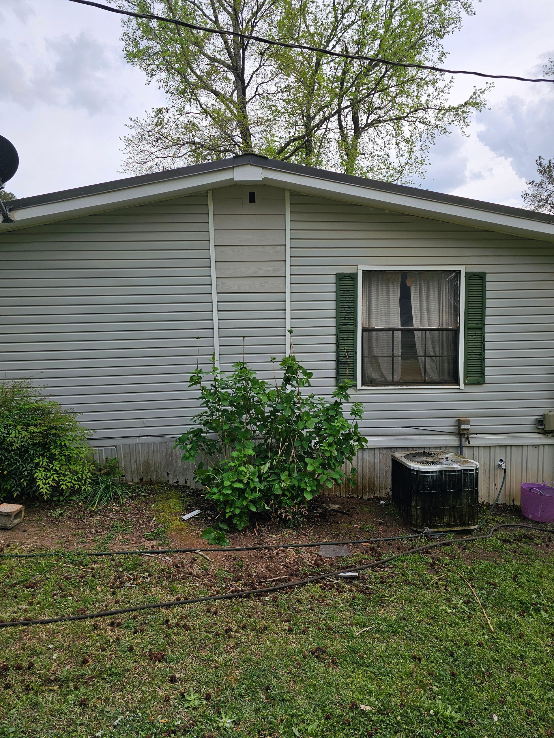Side of a light gray house with a window, green bushes, and an old air conditioner.
