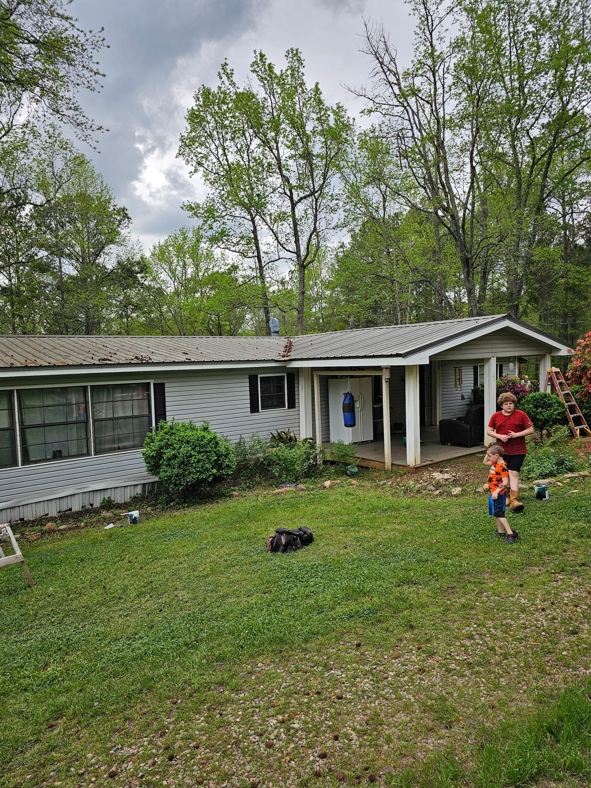 A single-story house with two children running on the front lawn. Green grass, trees, and cloudy sky.