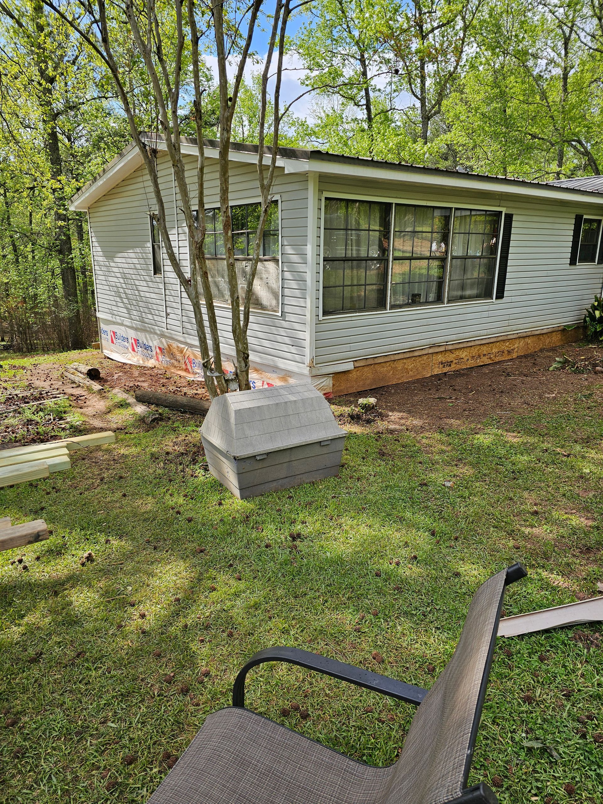 Small white building with multiple windows in a grassy yard, a tree in front, and a lounge chair in the foreground.