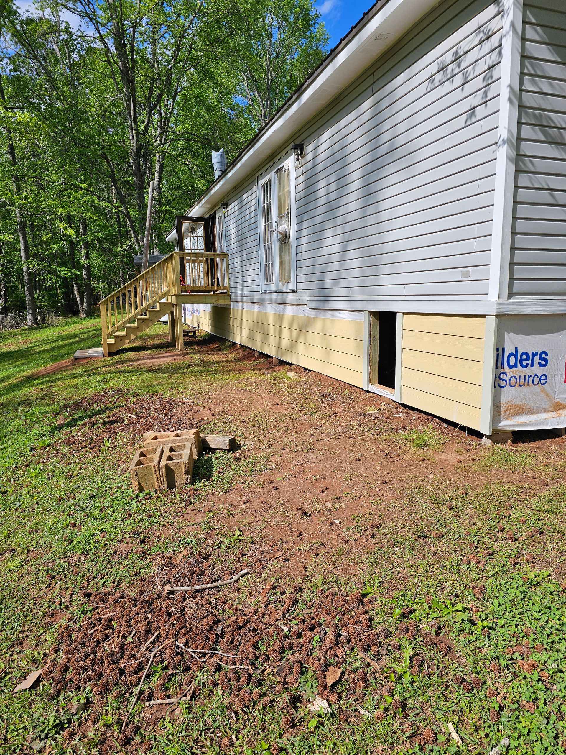 Side view of a house with light gray siding, wooden deck, and exposed foundation. Beige paneling and bare earth visible.