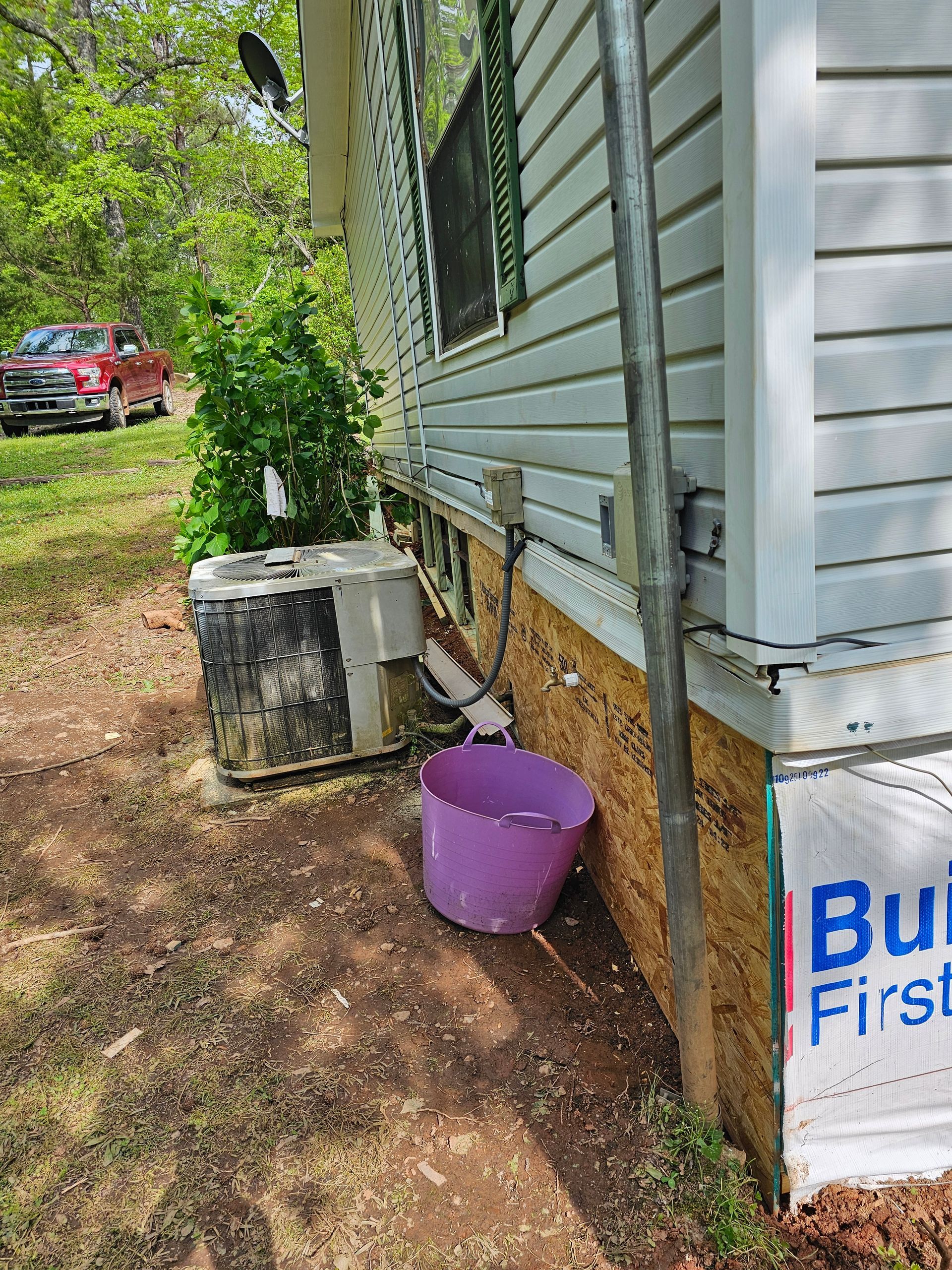 Side of a house with AC unit, purple bucket, and electrical box. Green siding, brown trim, and a car in the distance.