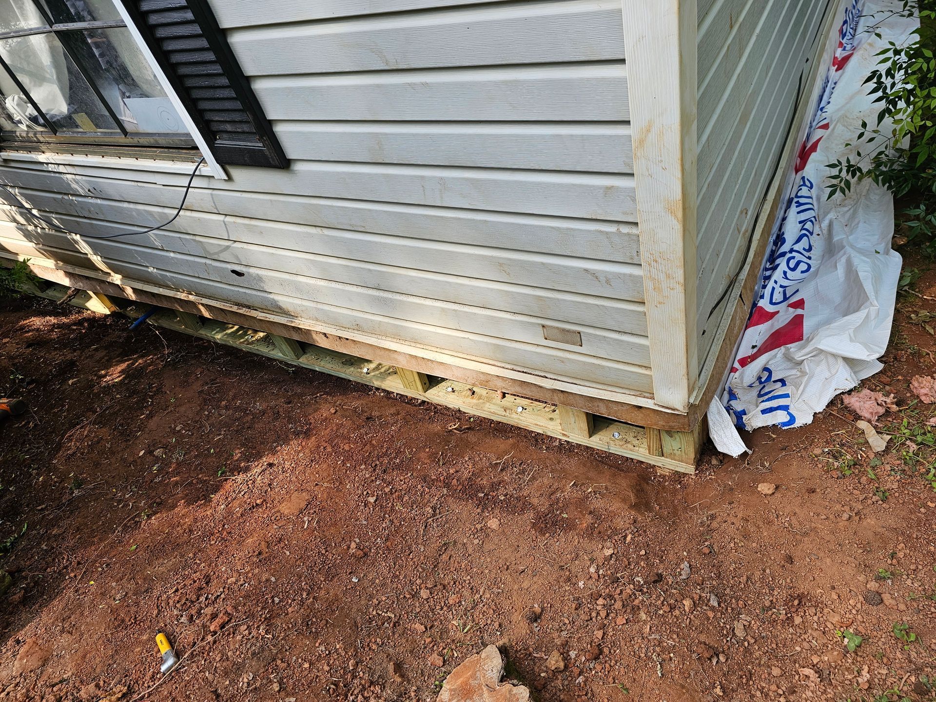 Close-up view of a building's siding and base. Light gray siding on wooden framing, resting on reddish-brown soil.