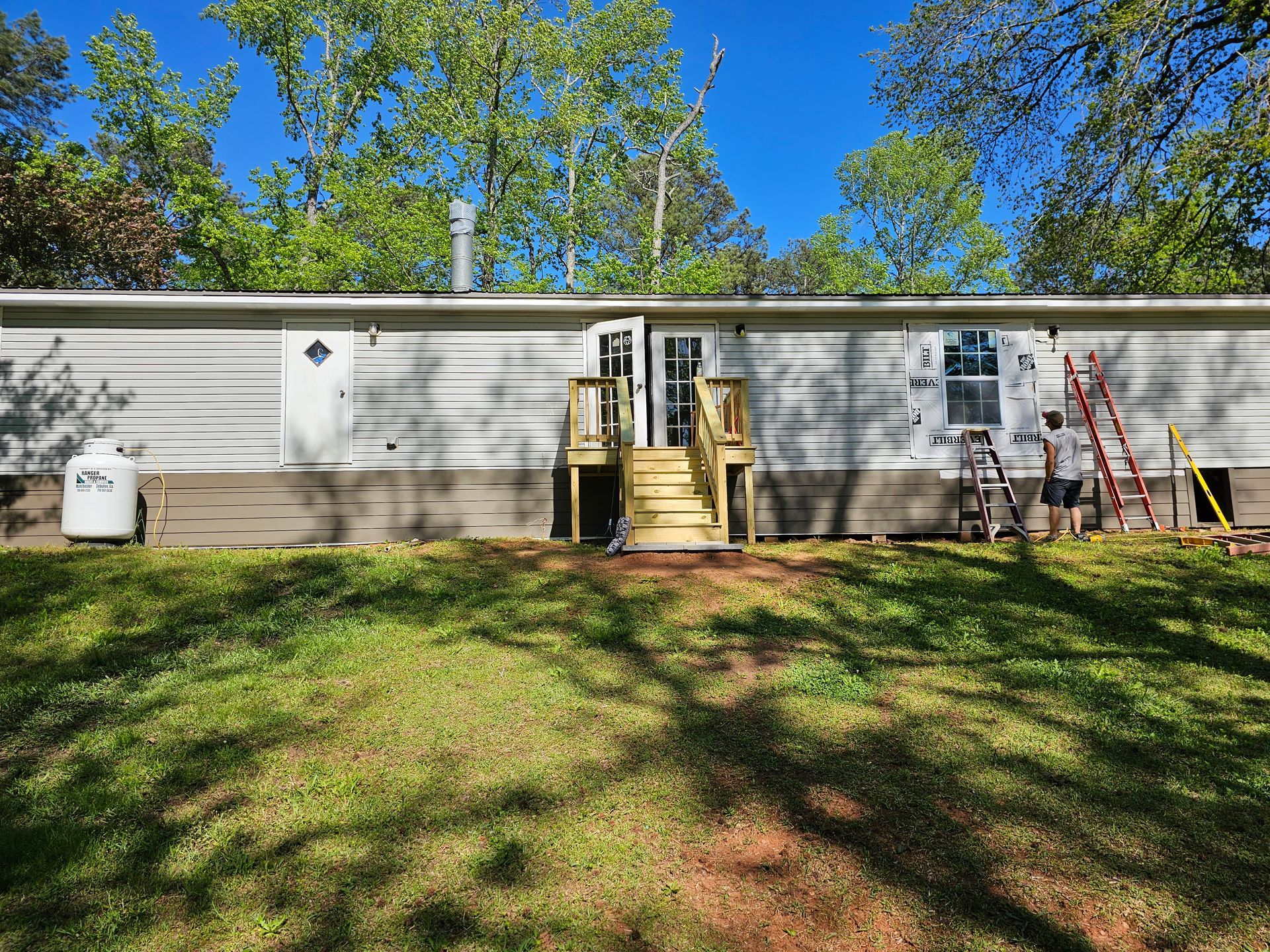 Mobile home with exterior doors, steps, and a propane tank, set in a grassy yard under a blue sky.