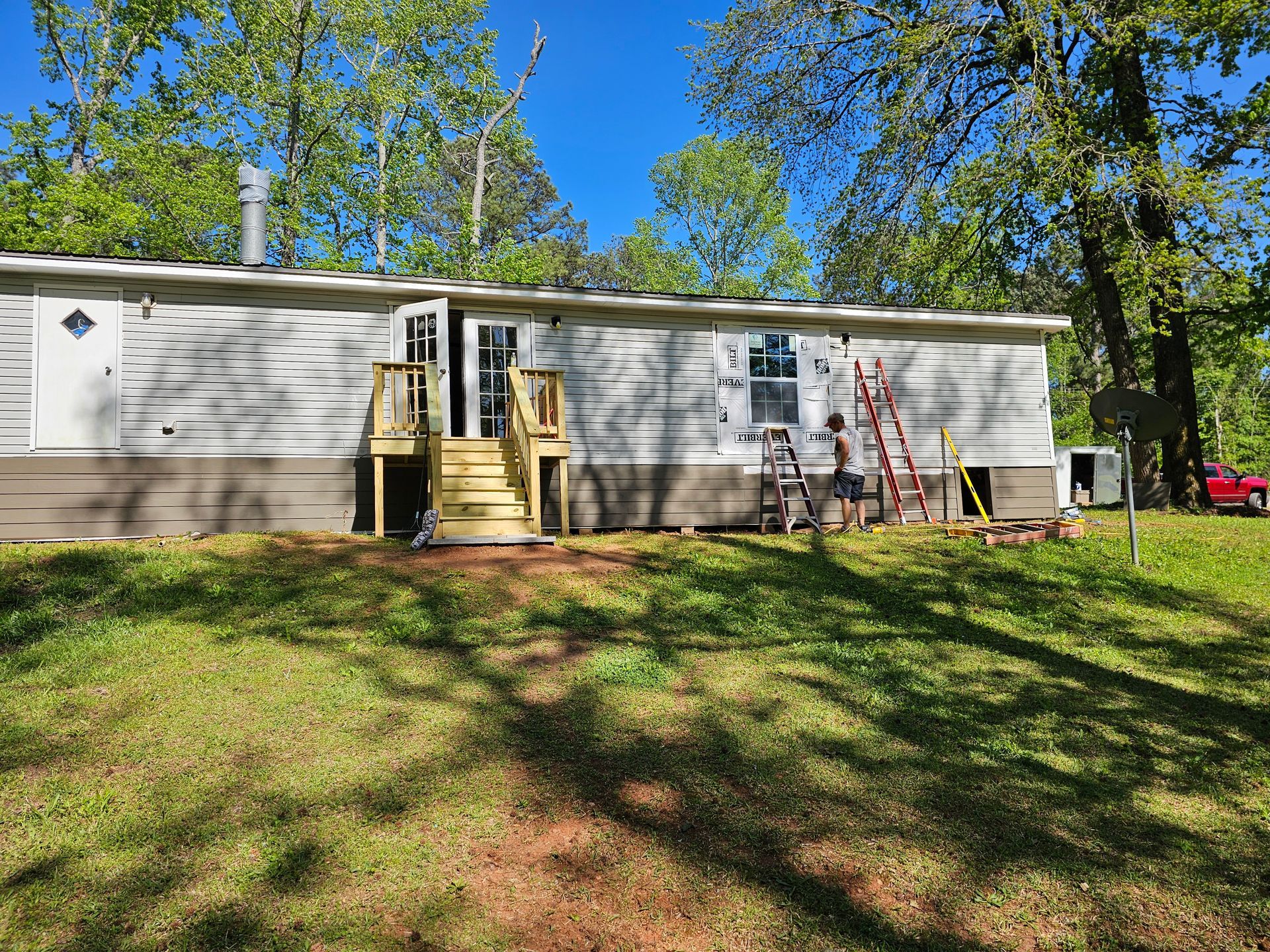 Mobile home with wooden stairs, green grass, and trees. A red truck is parked nearby.