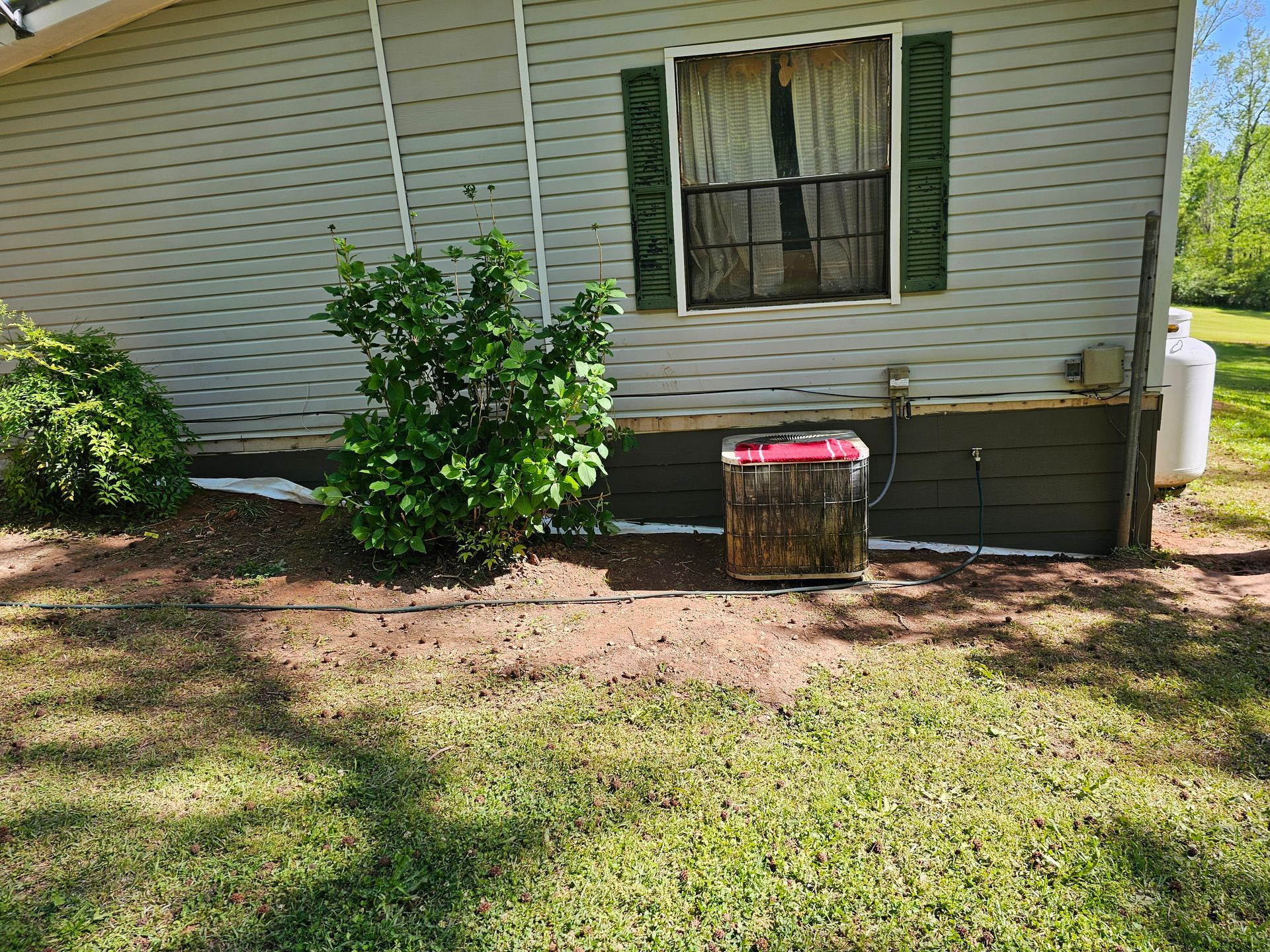 A house exterior with green siding, a window, and a propane tank, surrounded by a lawn and shrubs.