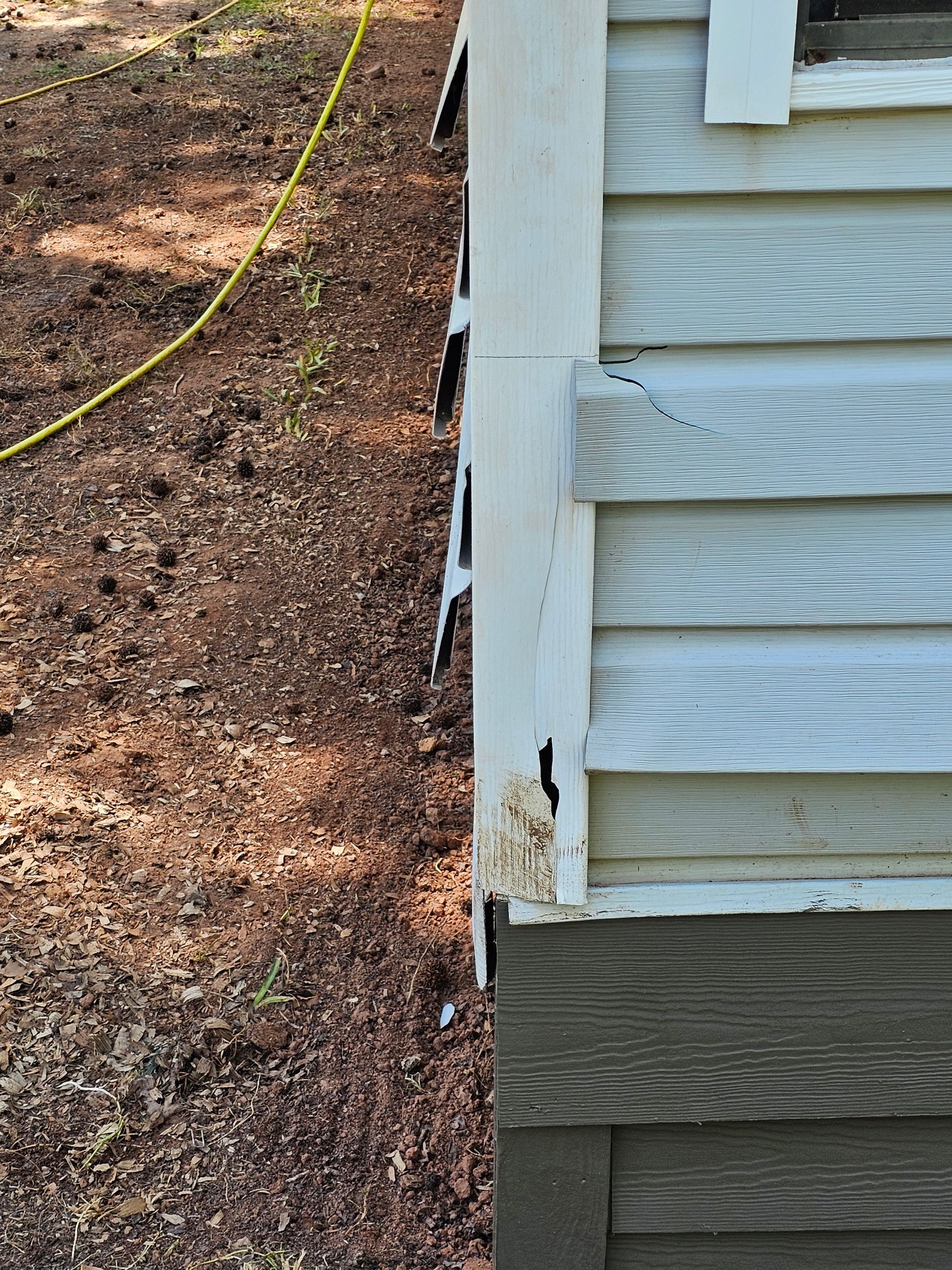 Exterior corner of a building with white siding, showing deterioration and damage, next to a brown dirt patch.