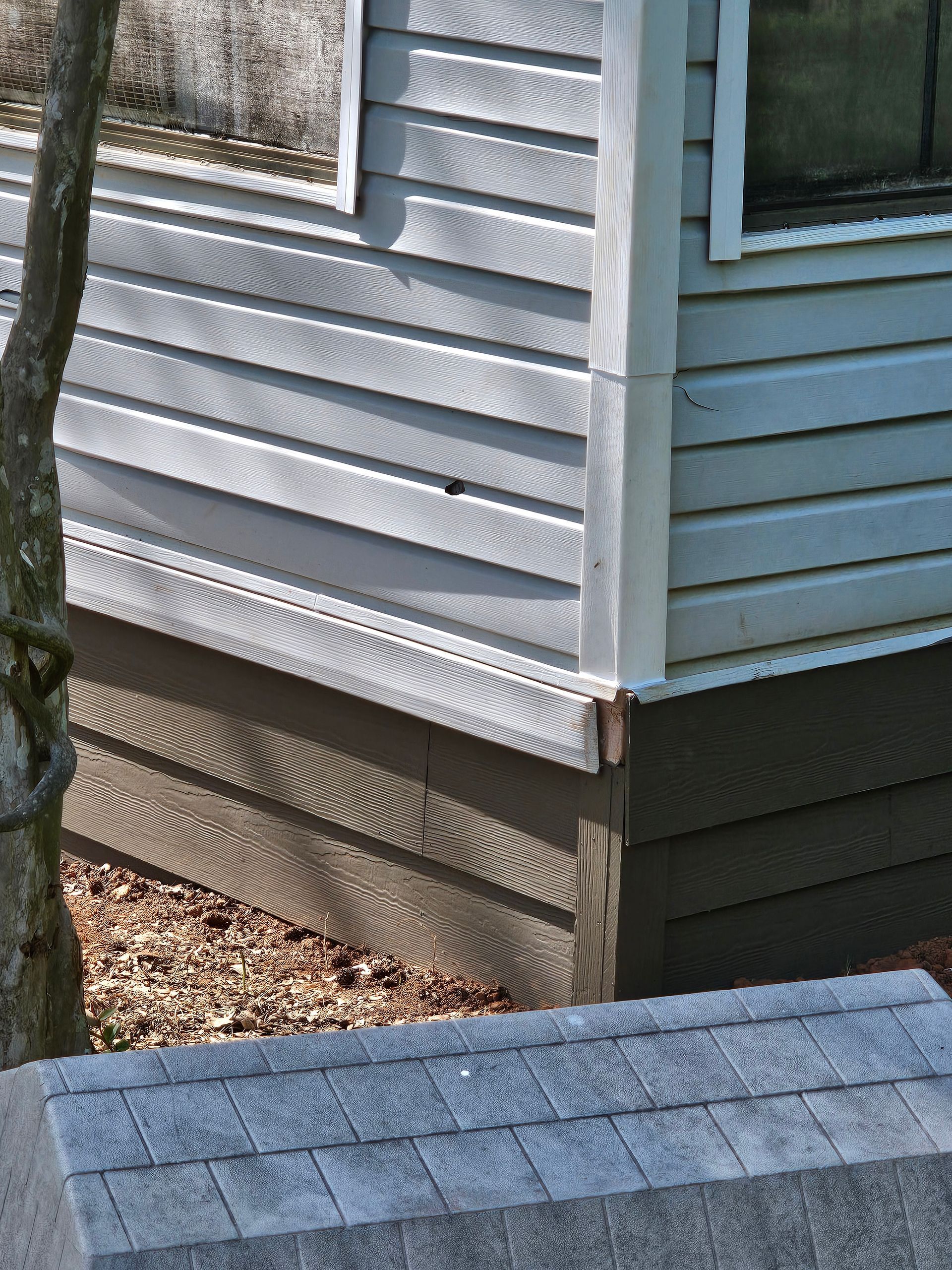 Corner of a light blue house with horizontal siding. Brown foundation and a portion of a gray roof visible.