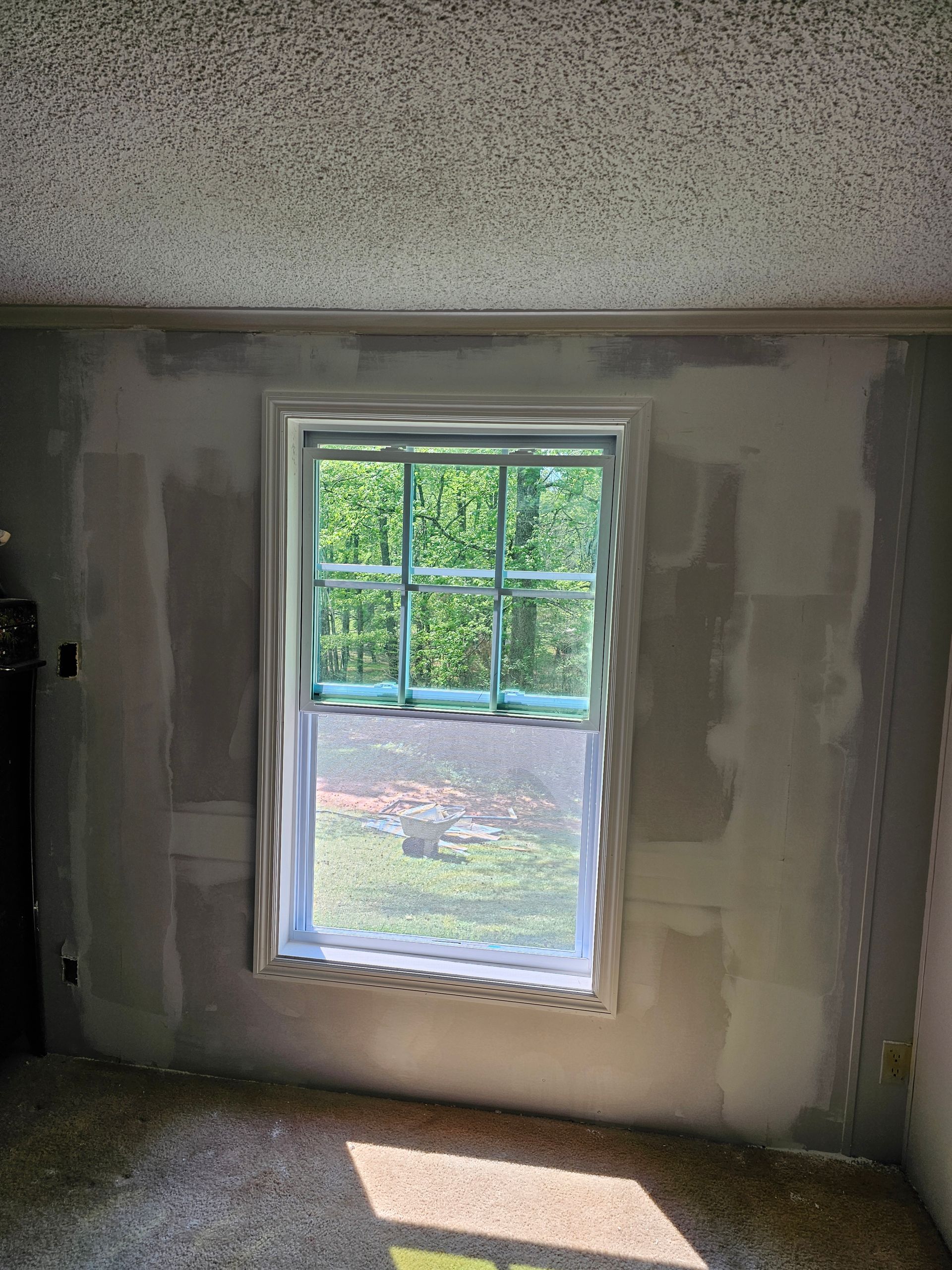 Window in a room with unfinished drywall, looking out onto trees. Sunlight streams in.