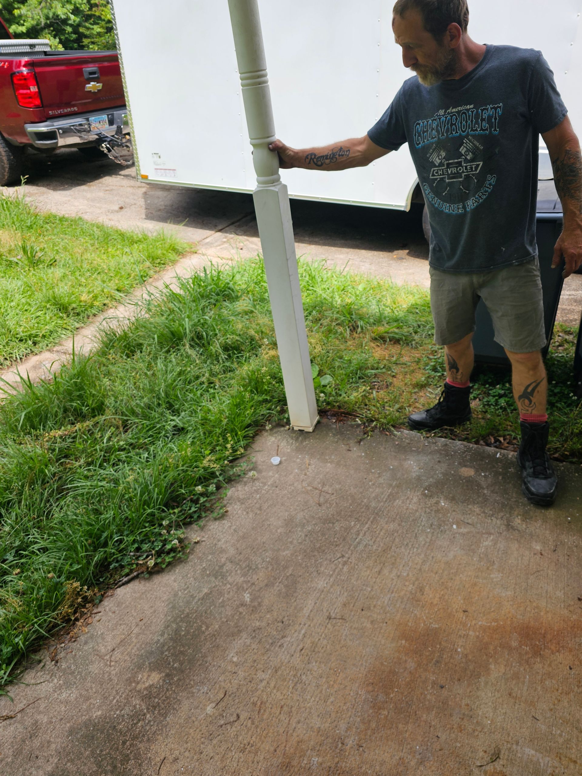 Man standing by a white support column on a concrete surface, with a truck and trailer in the background.