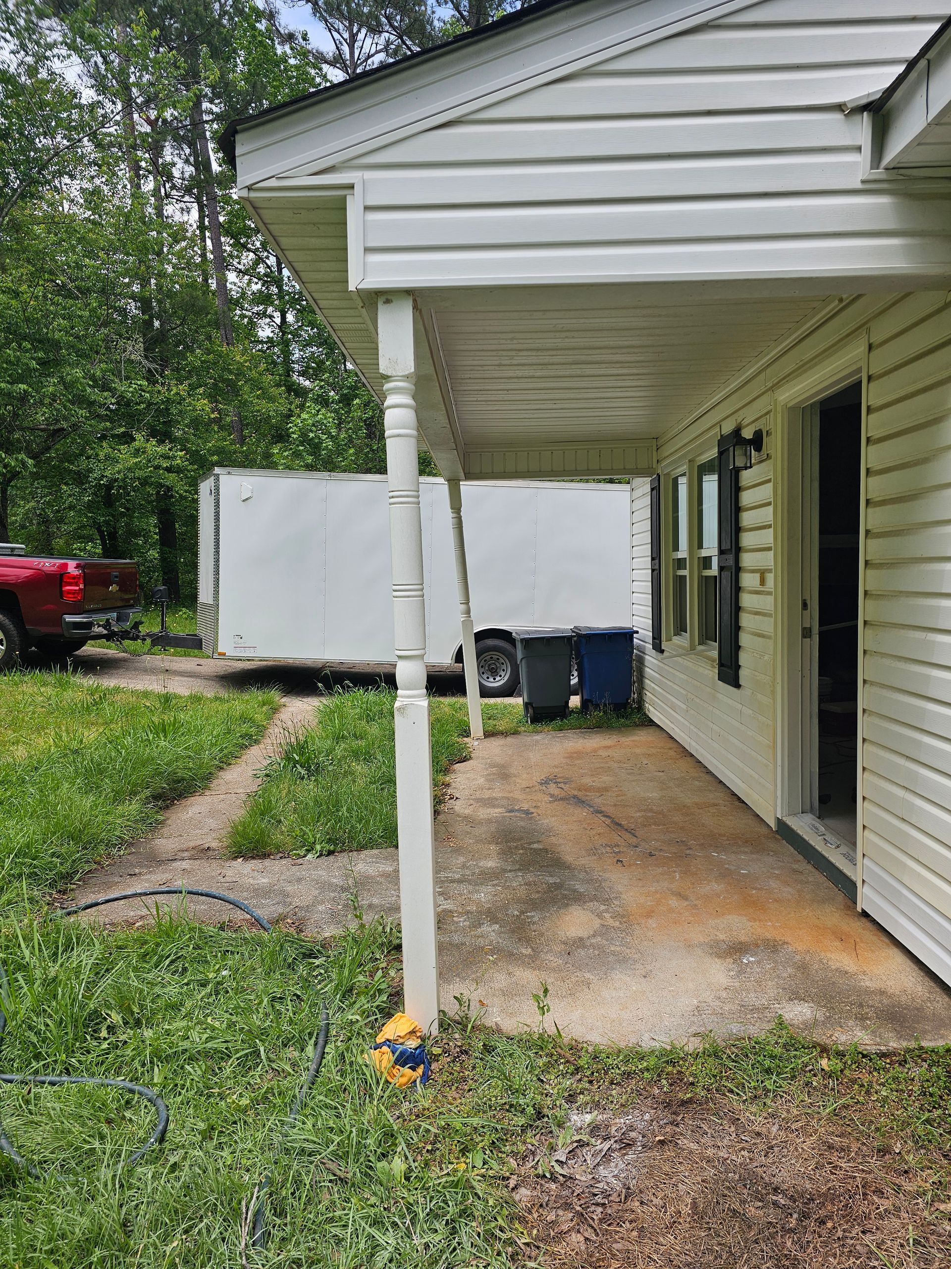 White house porch with concrete slab, white columns, and trailer in the background. Green grass and blue trash bins.