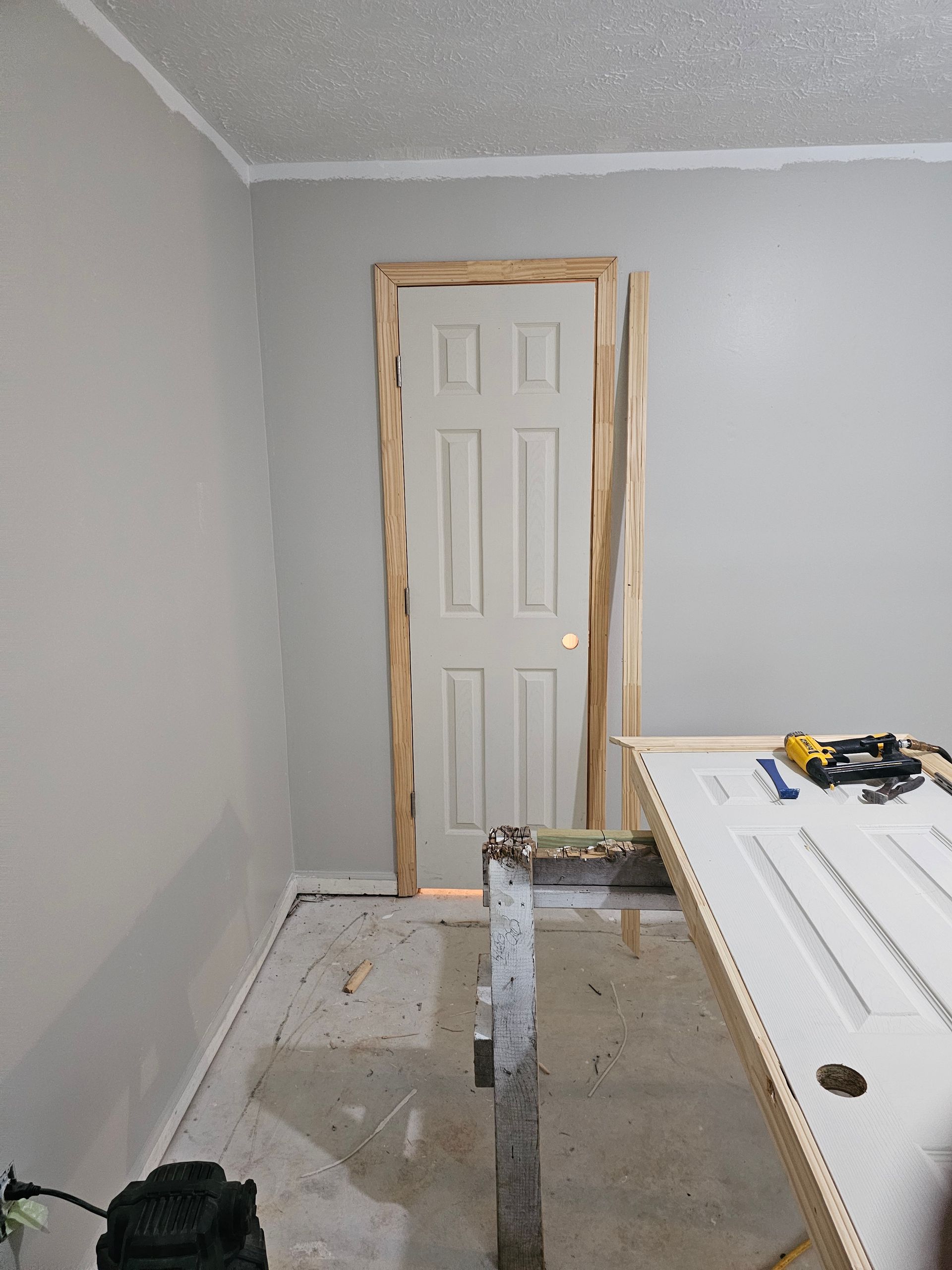 Interior room with a white door, light gray walls, and unfinished trim. A workbench with tools is in the foreground.