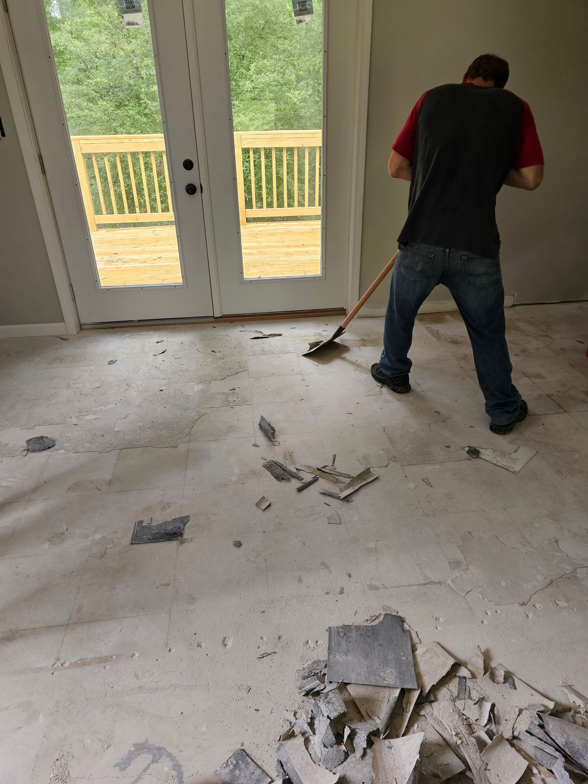 A person is scraping debris off a concrete floor, French doors and a deck in background.
