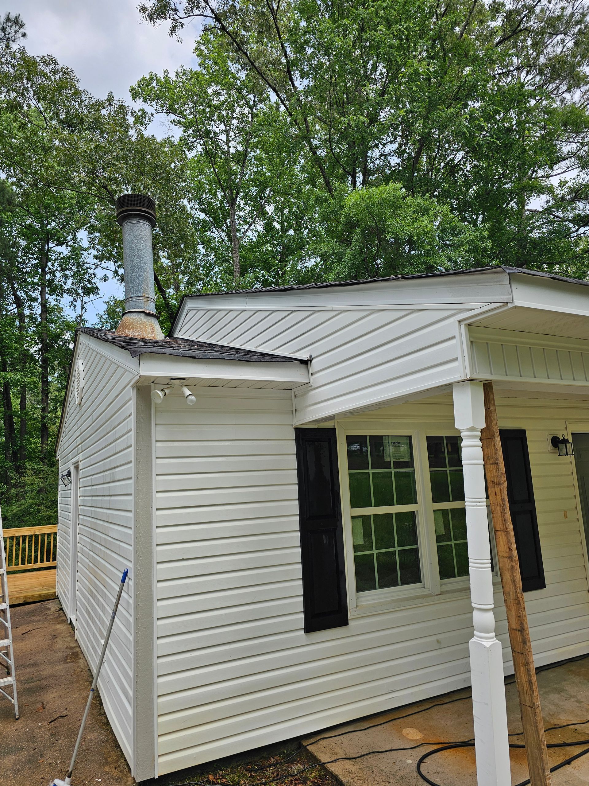 White house with chimney, black shutters, porch, siding, and surrounded by trees.