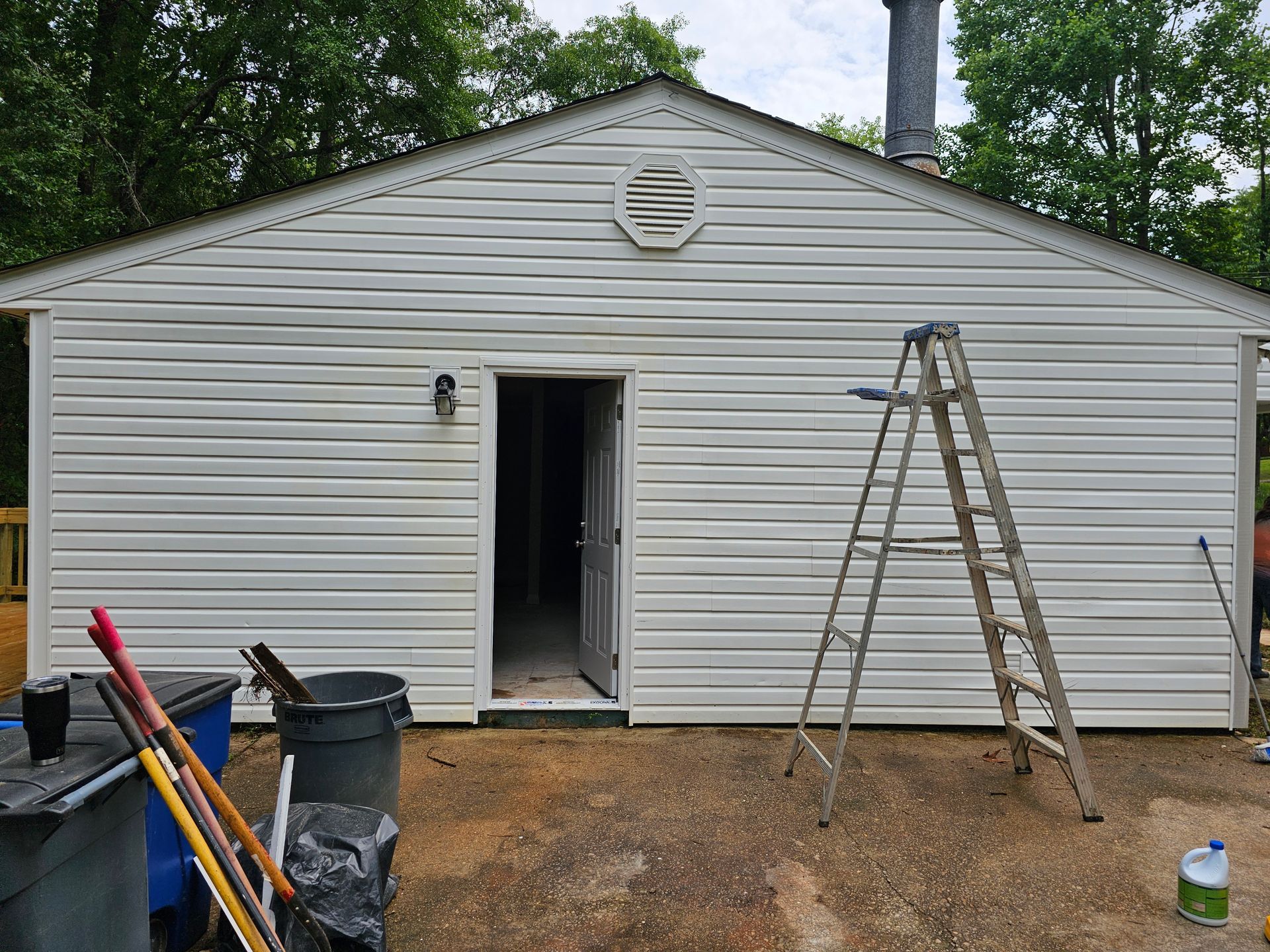 White-sided building with open door, vent, chimney, ladder, and trash cans on a gravel surface.