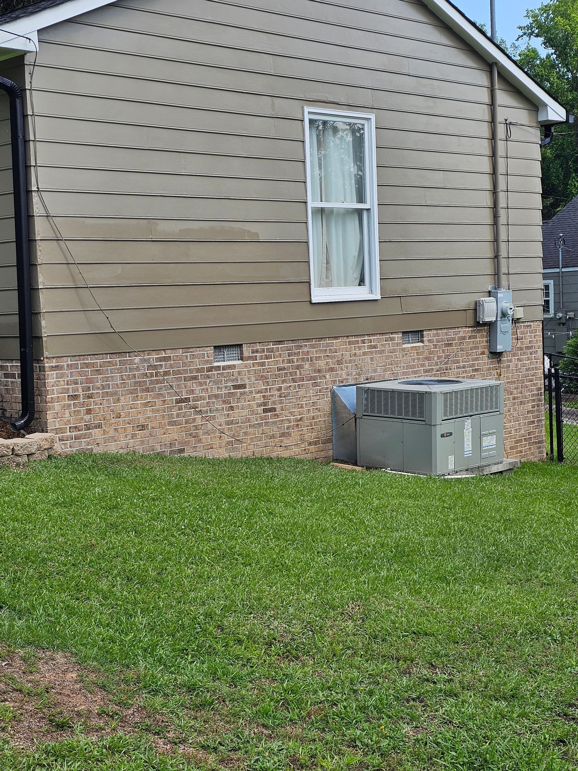 Side of a house with tan siding and brick base, a window, and an air conditioning unit on green grass.