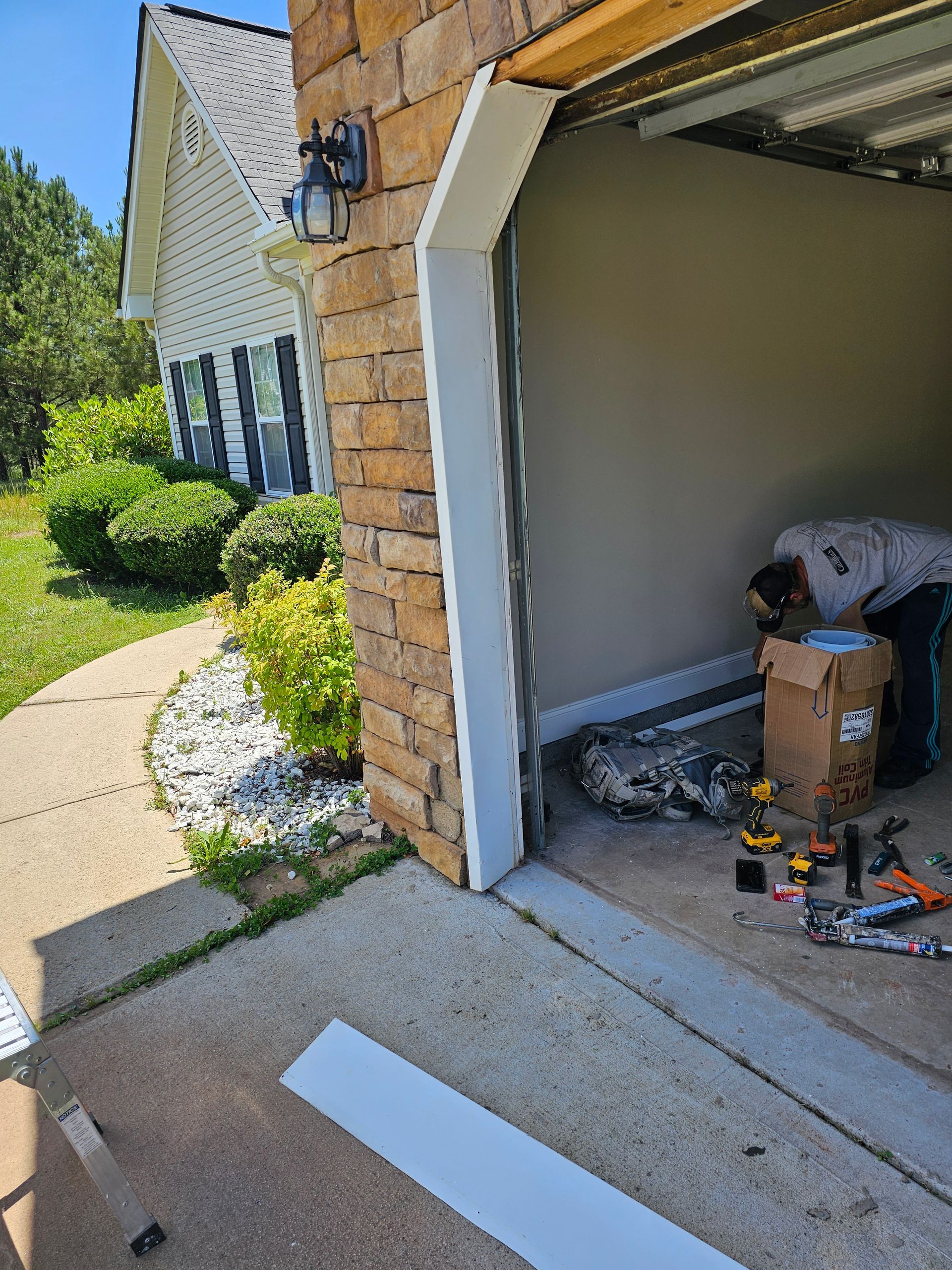 Person repairing garage door frame on a sunny day. White trim, brick facade, and sidewalk.