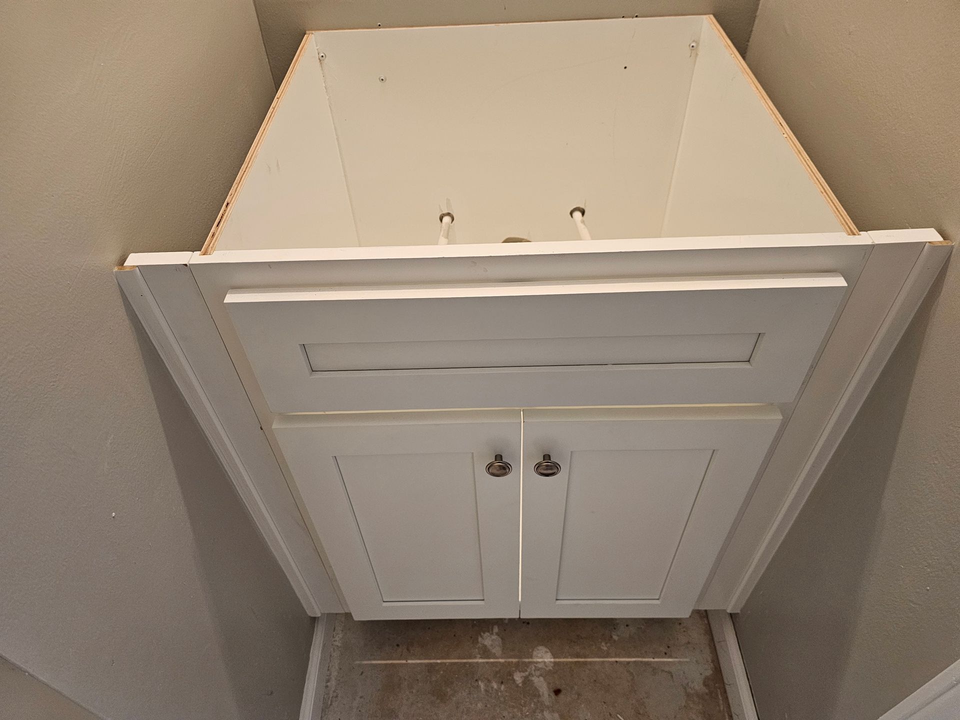 White bathroom cabinet installed in a corner between beige walls. Two doors below a drawer.