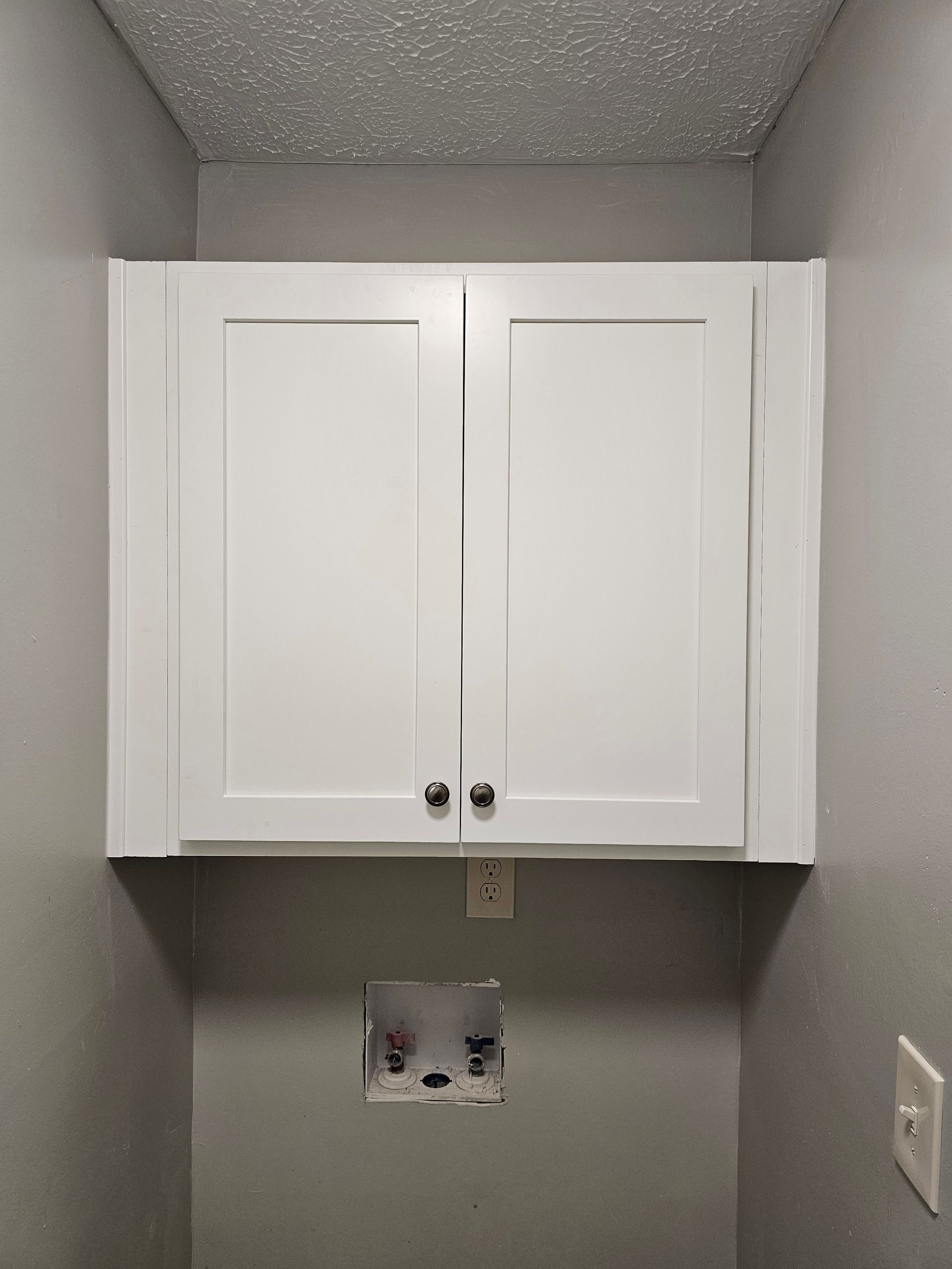 White cabinet above laundry hookups in a gray-painted laundry room.