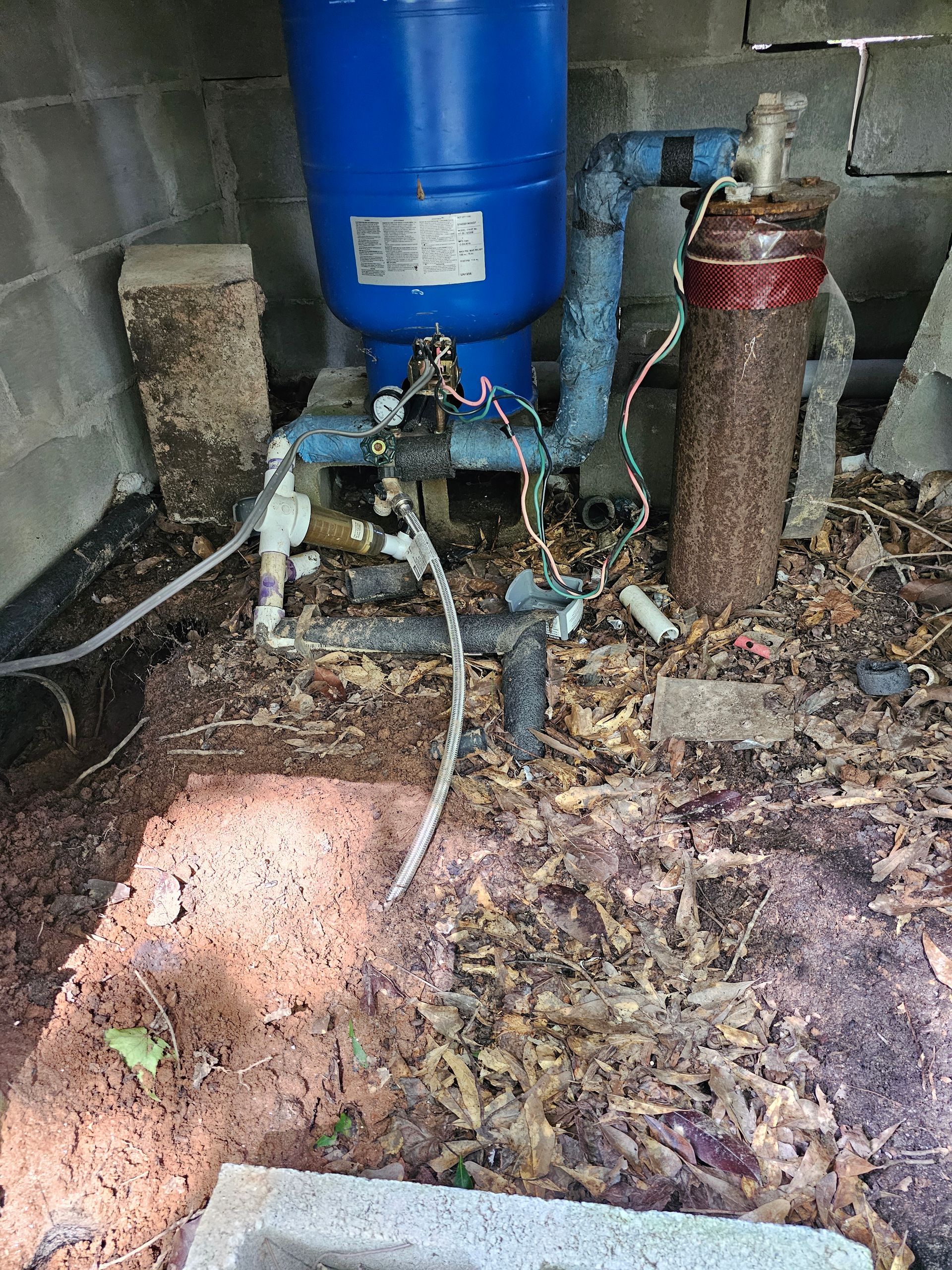 Blue water pressure tank with connected pipes and filters in a debris-filled area.