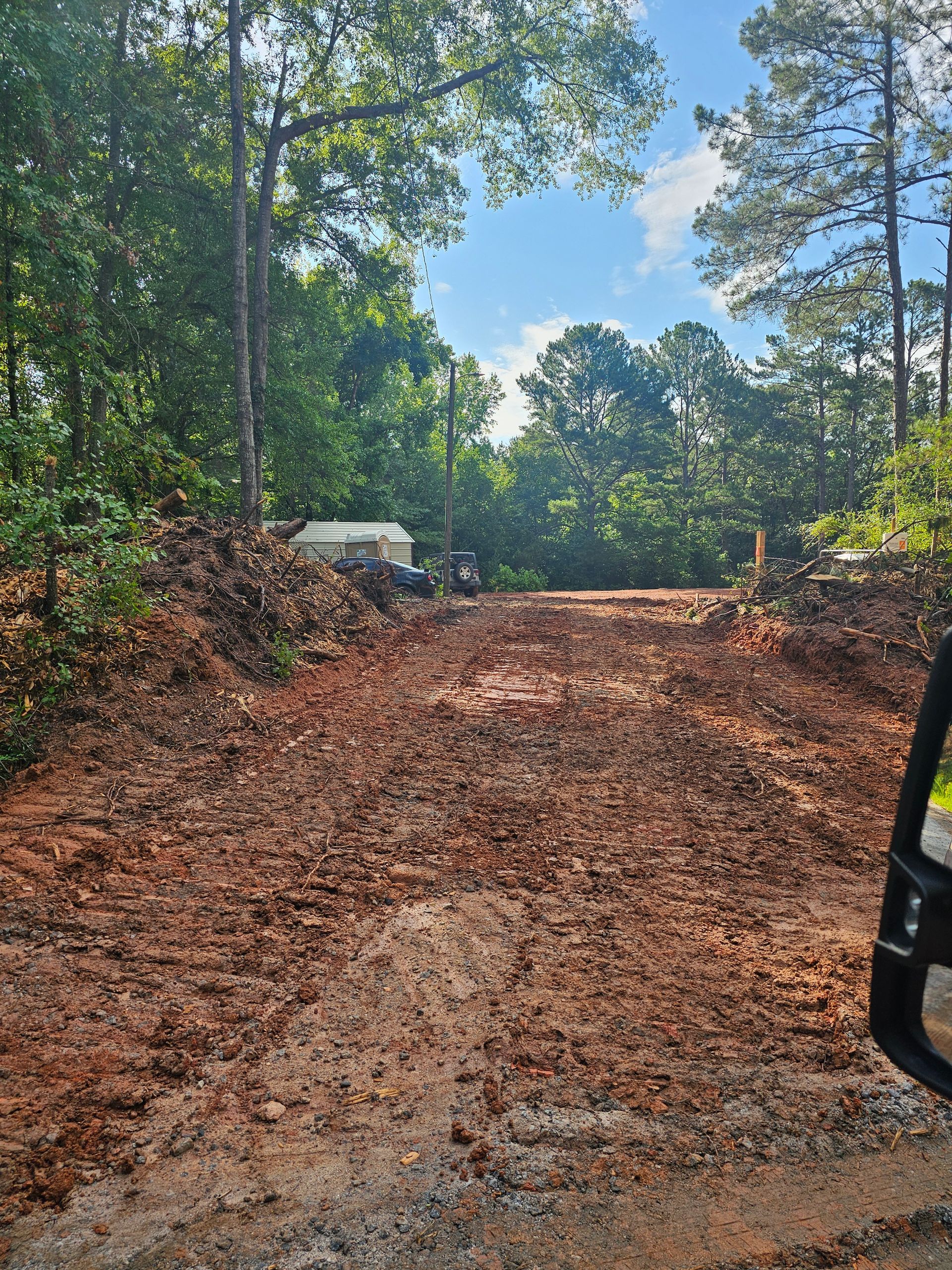A dirt road leads through a wooded area. The road is red clay and appears recently graded.