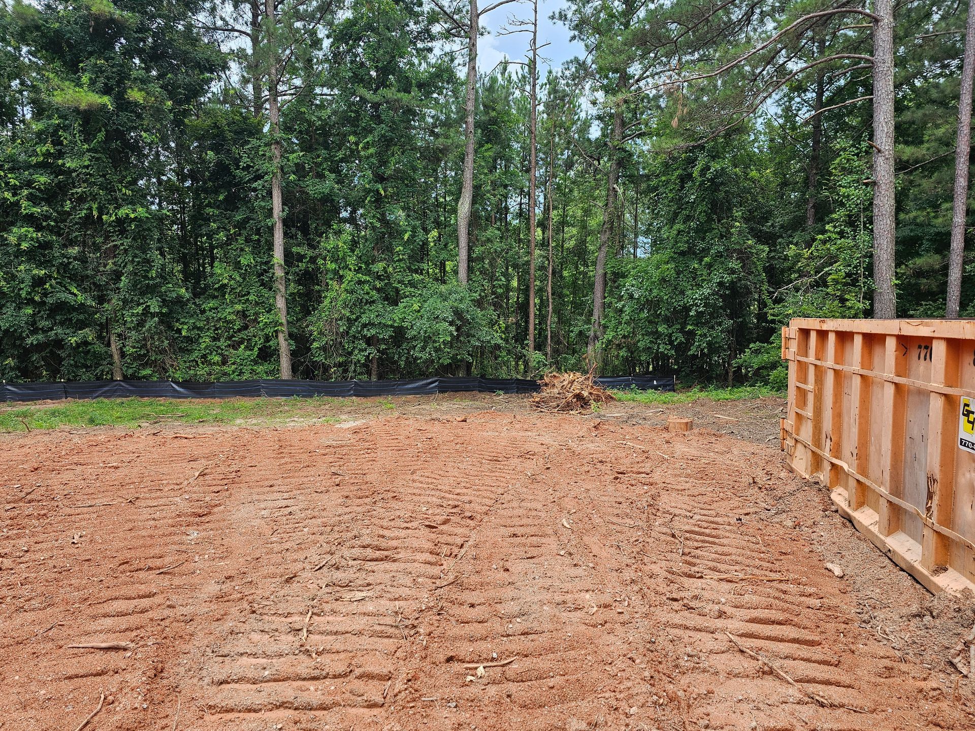 Cleared red soil area with trees in the background, a wooden structure on the right.