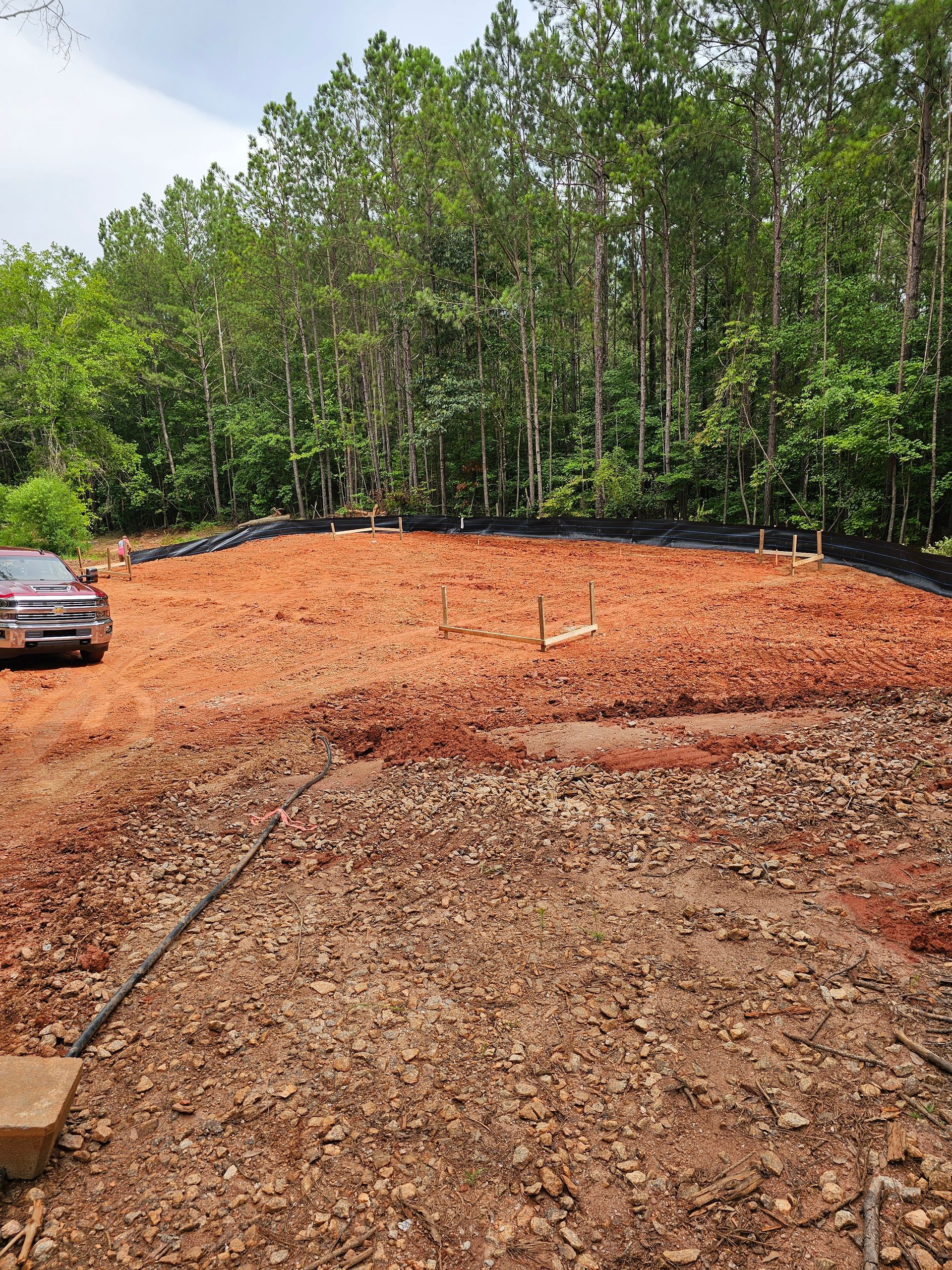 Cleared construction site with red soil, surrounded by trees and a vehicle.