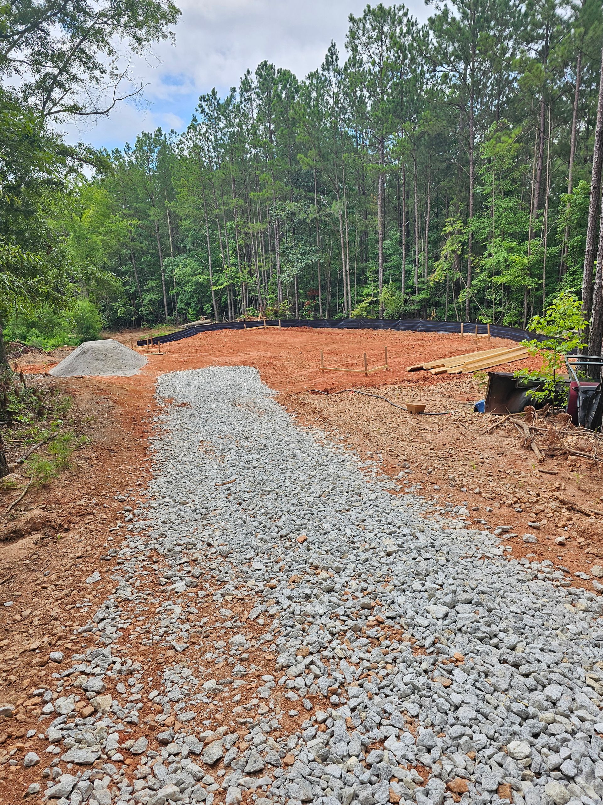 Gravel pathway leads to a cleared construction site in a wooded area with trees in the background.